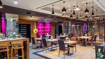 A modern restaurant interior with multiple dining tables, chairs, pendant lighting, and a vibrant wall decoration.