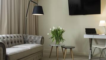 A modern living room with a tufted sofa, a flat-screen TV, a desk with a laptop, a lamp, and a vase with white flowers.