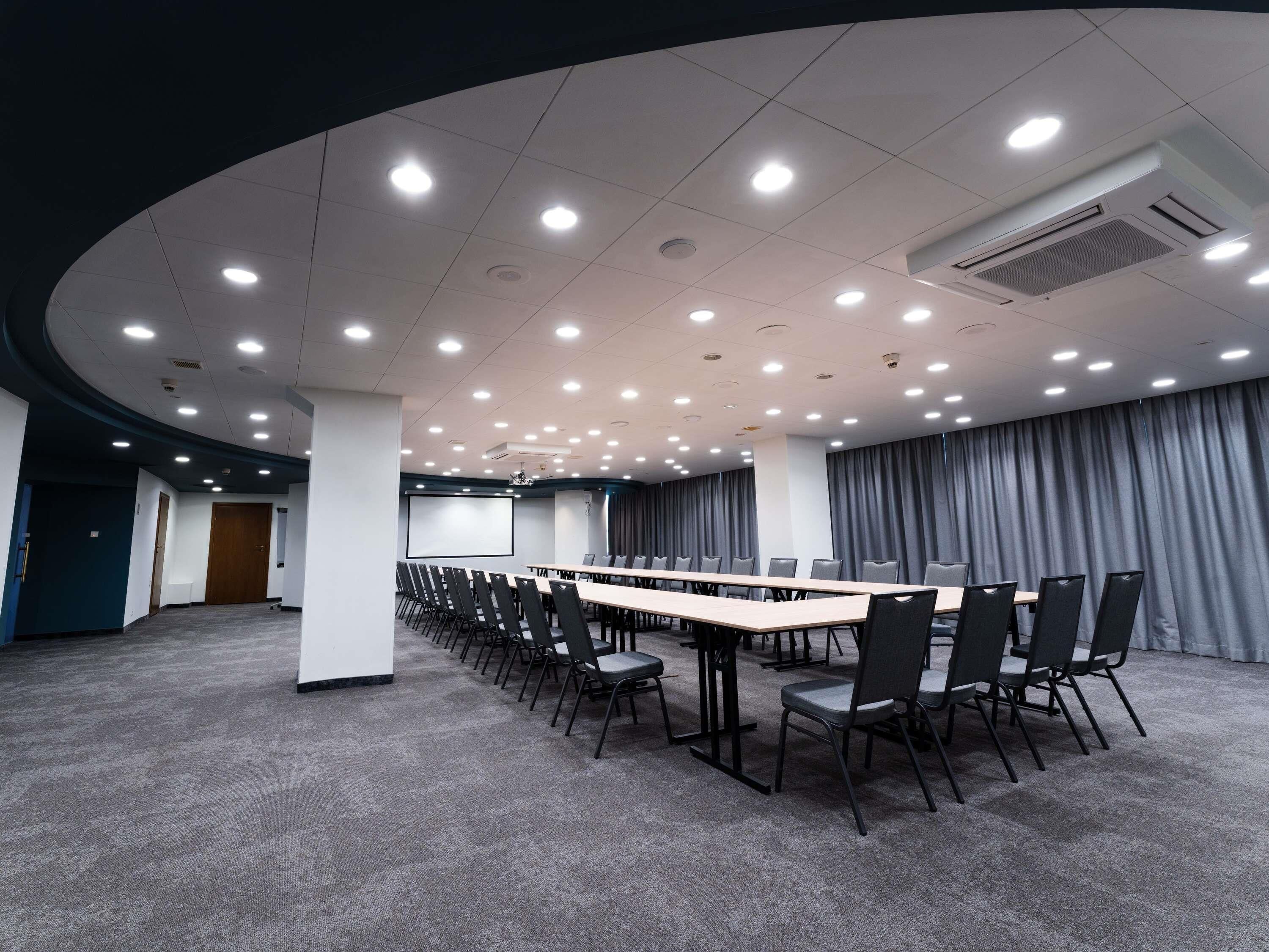 Ein Konferenzraum mit einem langen Tisch, Stühlen, einer Projektionsleinwand und einem grauen Vorhang im Hintergrund.