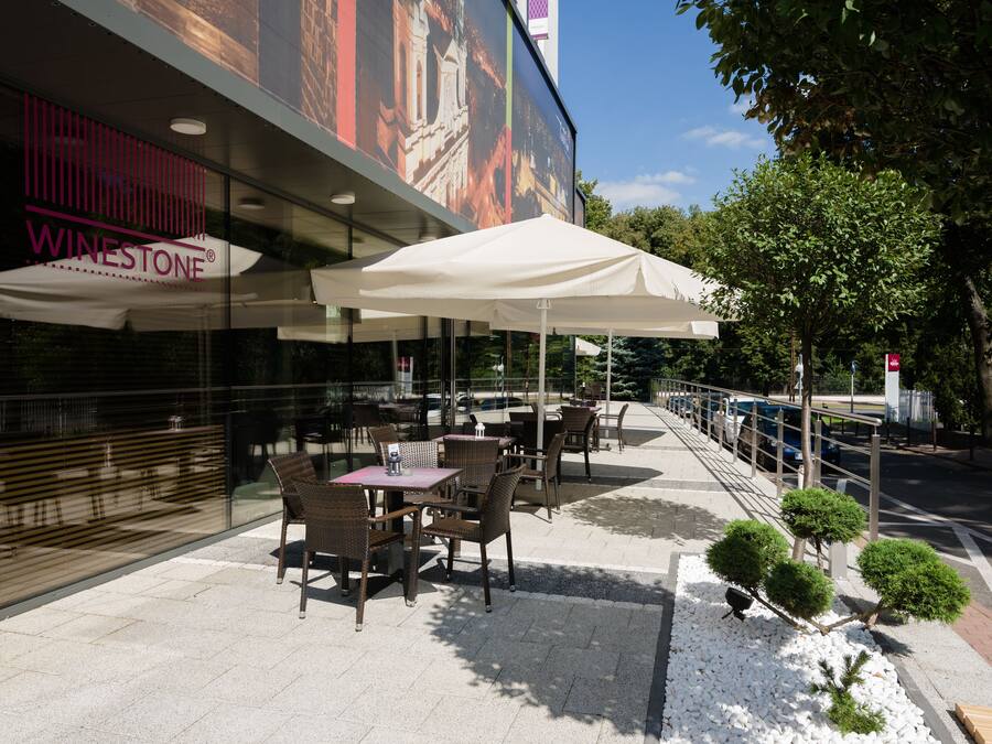 An outdoor seating area with tables and chairs, a large mural on the building, and a white umbrella providing shade.
