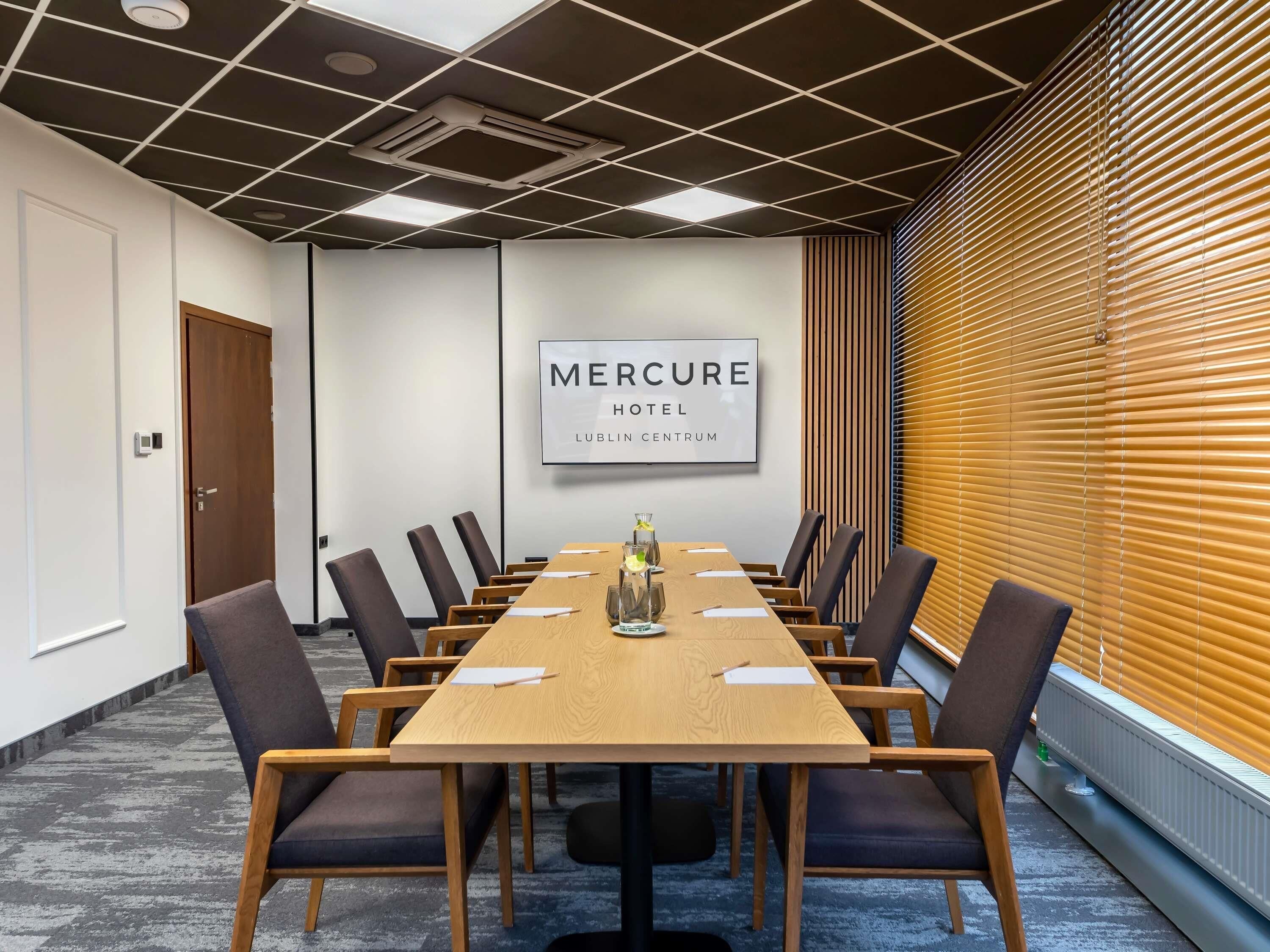 Un salón de conferencias con una larga mesa, sillas, un cartel enmarcado y una ventana con persianas.