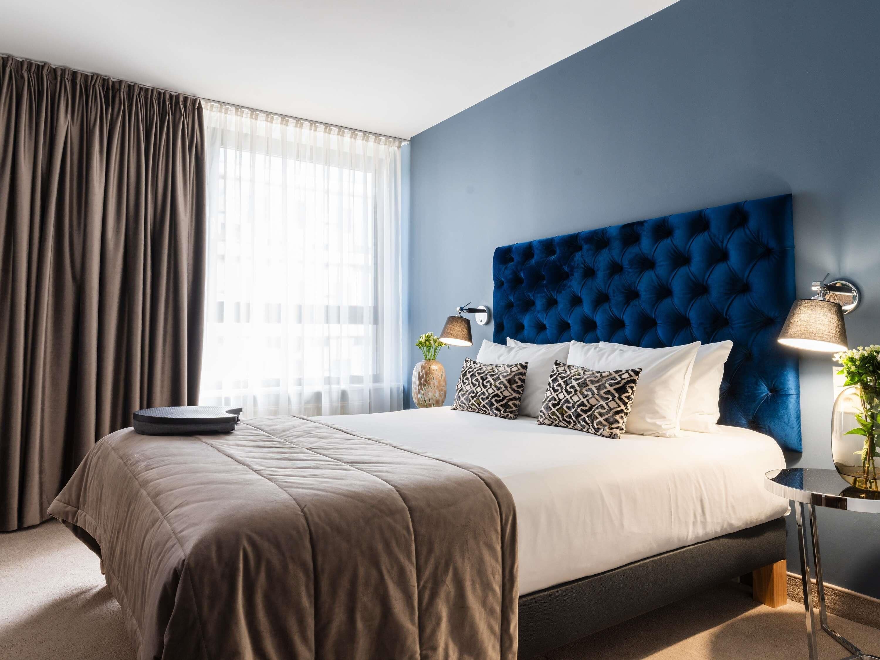 Una habitación de hotel moderna con una cama grande, cabecera, mesita de noche con lámpara y una ventana con cortinas.