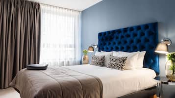Una habitación de hotel moderna con una cama grande, cabecera, mesita de noche con lámpara y una ventana con cortinas.