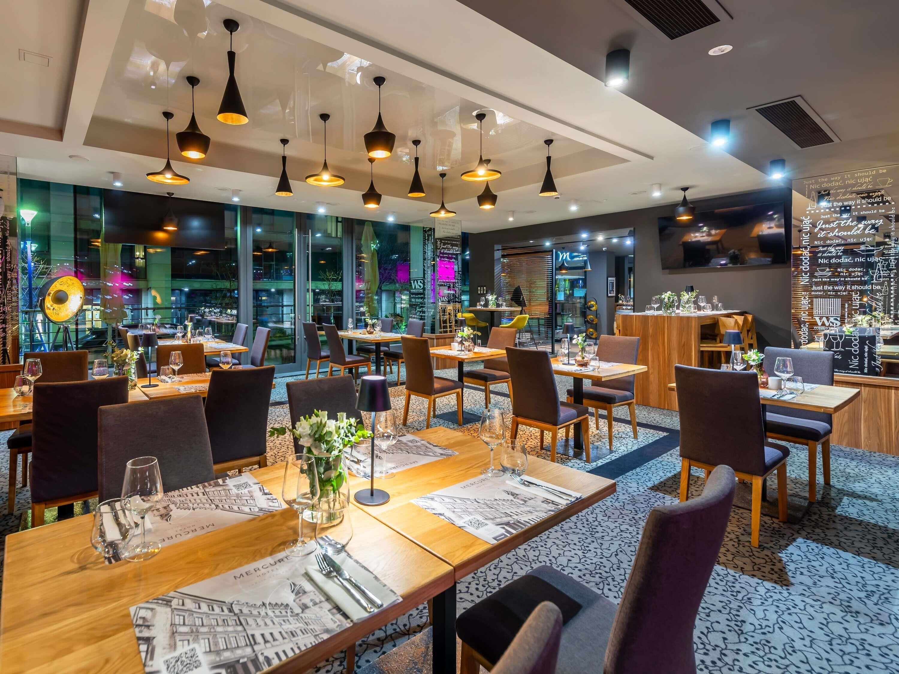 Un restaurant moderne doté de nombreuses tables, d’un éclairage suspendu noir et offrant une vue sur le paysage urbain grâce à de grandes fenêtres.