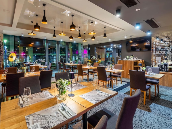 A modern restaurant with multiple dining tables, black pendant lighting, and a view of the cityscape through large windows.