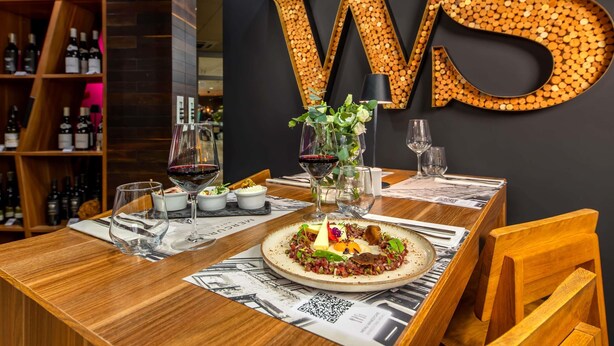 A restaurant interior with a wooden table, wine glasses, a decorative wall, and a bookshelf with wine bottles. - Hotel Mercure Lublin Centrum (Lublin)