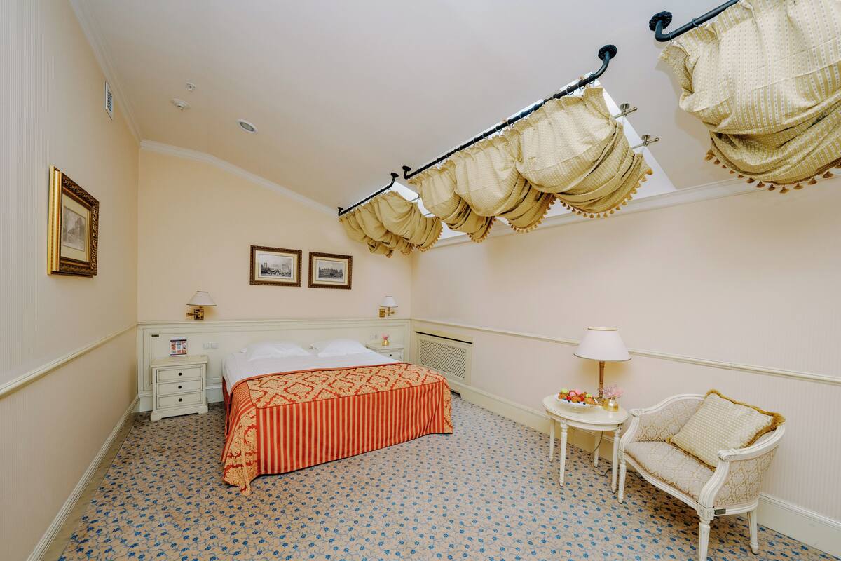 Un dormitorio con cama, mesita de noche, silla, pequeña mesa con lámpara y barra para cortinas en la pared.