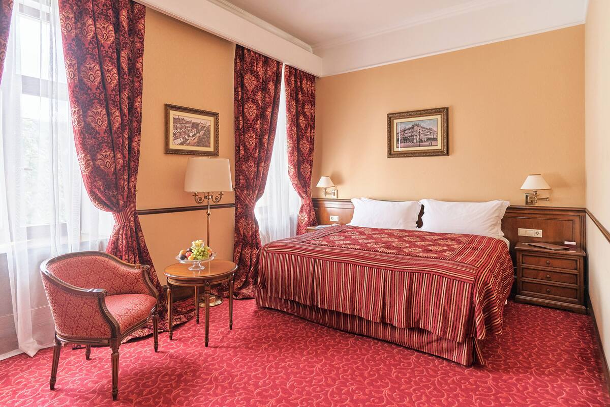 Habitación de hotel con una cama grande, una silla, una mesita de noche con una lámpara y dos cuadros en las paredes.