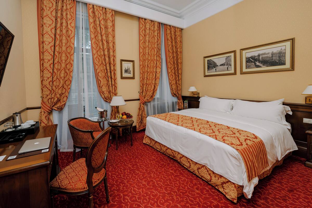 Habitación de hotel con una cama grande, un escritorio con una computadora portátil, una silla, una mesita de noche con una lámpara y dos cuadros en la pared.