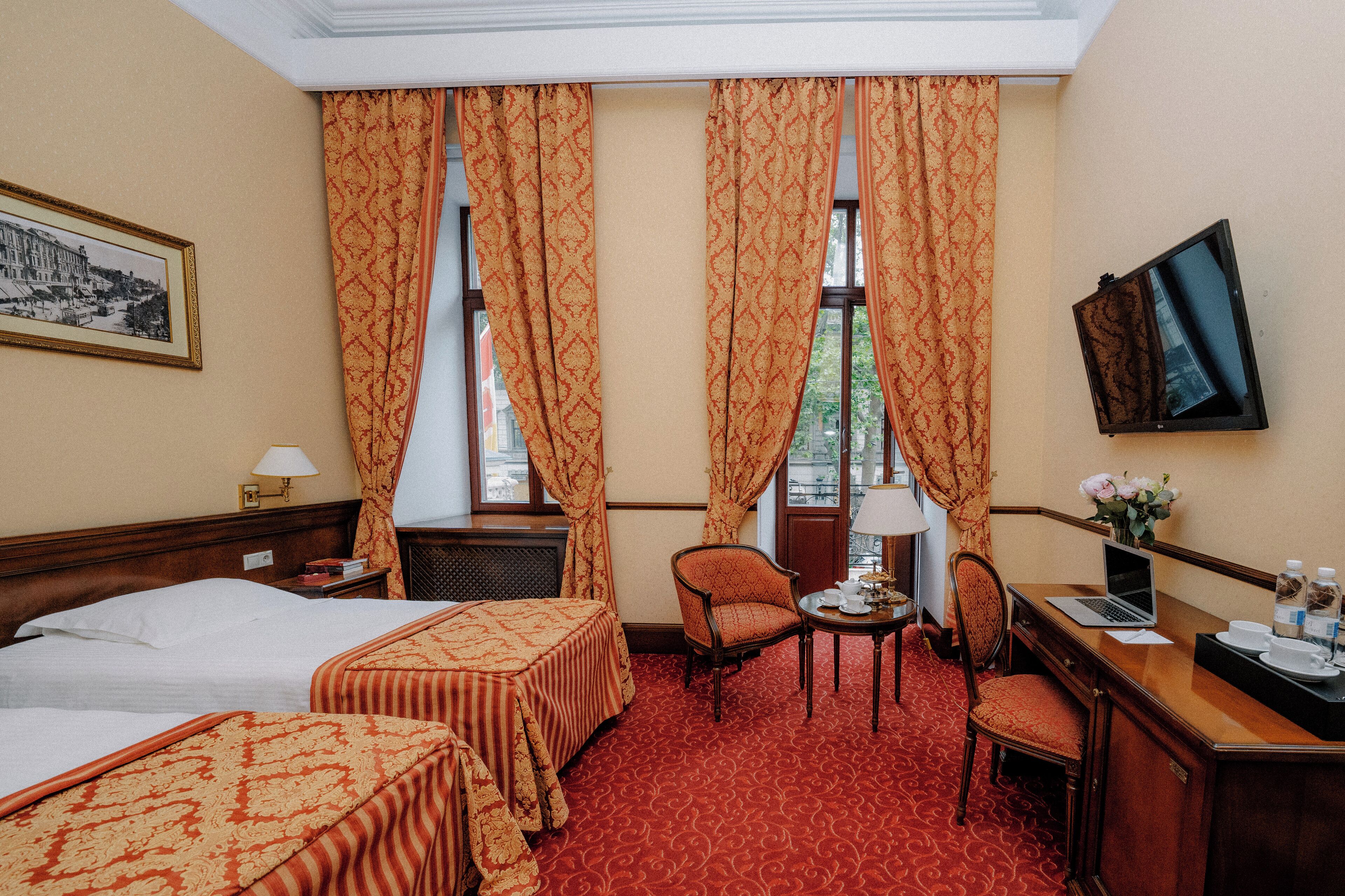 A hotel room with two beds, a desk with a laptop, a chair, a small table with a lamp, and a TV on the wall.