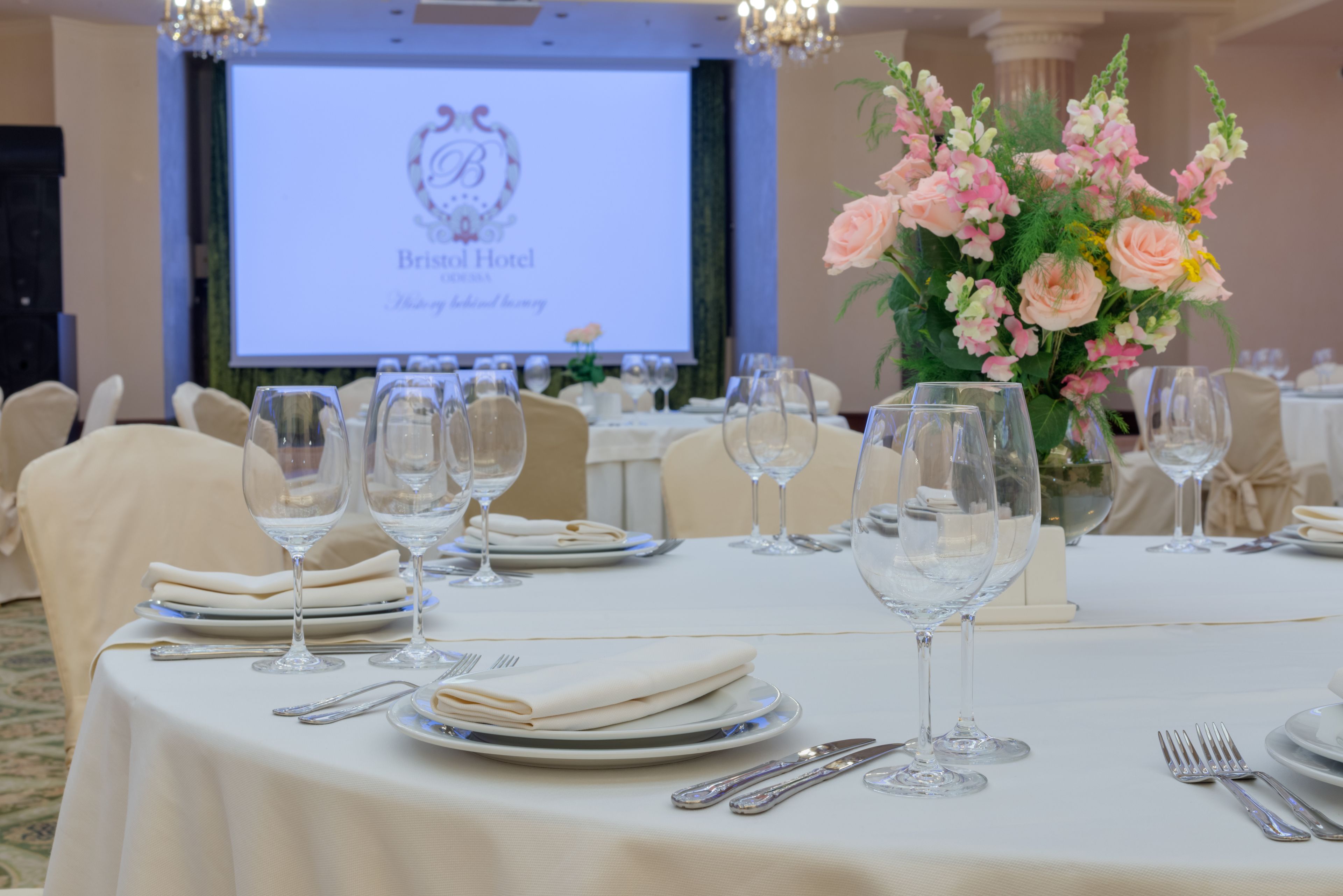 Una mesa de banquete dispuesta para un evento formal, con una pantalla de proyector que muestra el nombre del hotel al fondo.