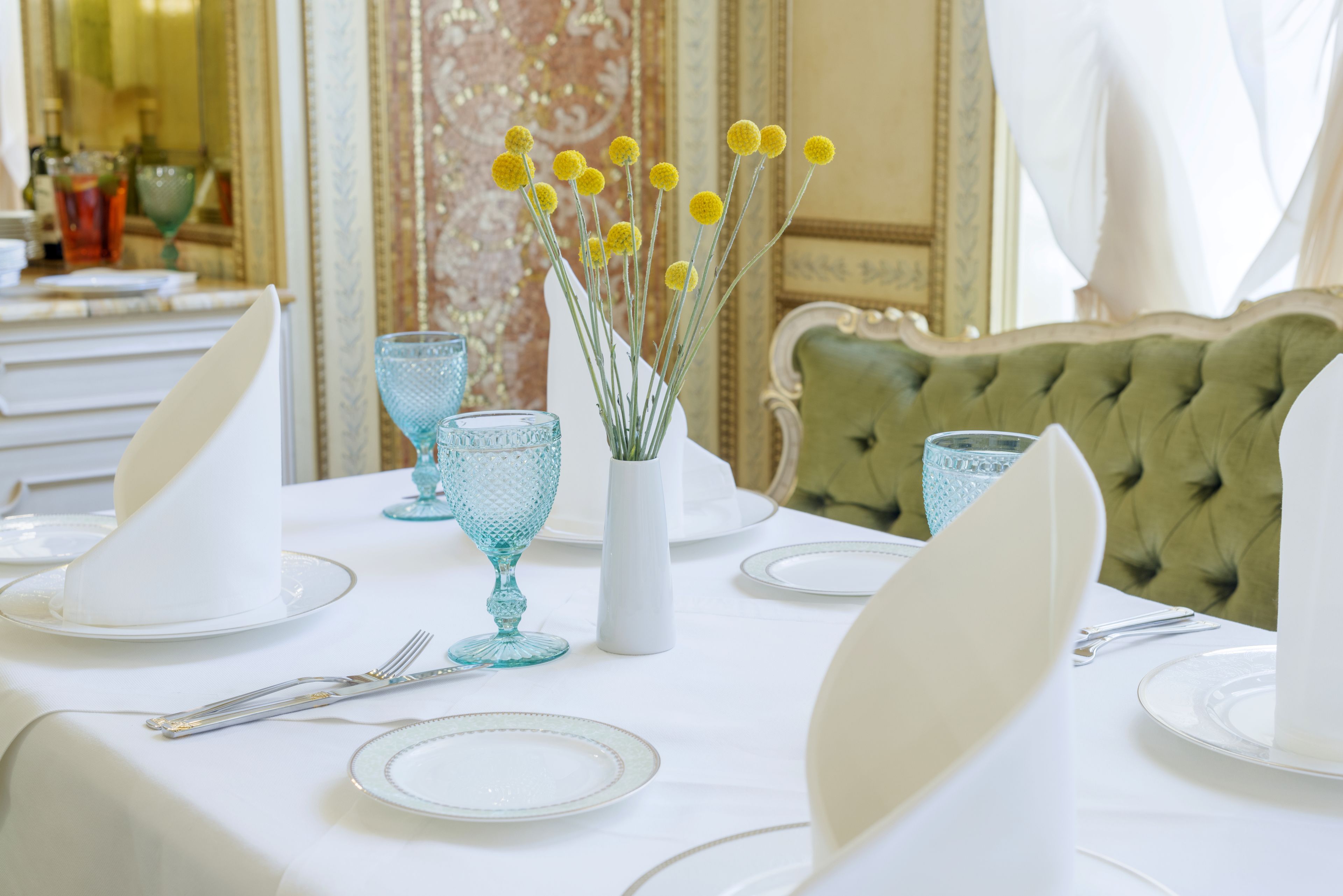 Una mesa de comedor servida con platos blancos, copas azules y un florero con flores amarillas.