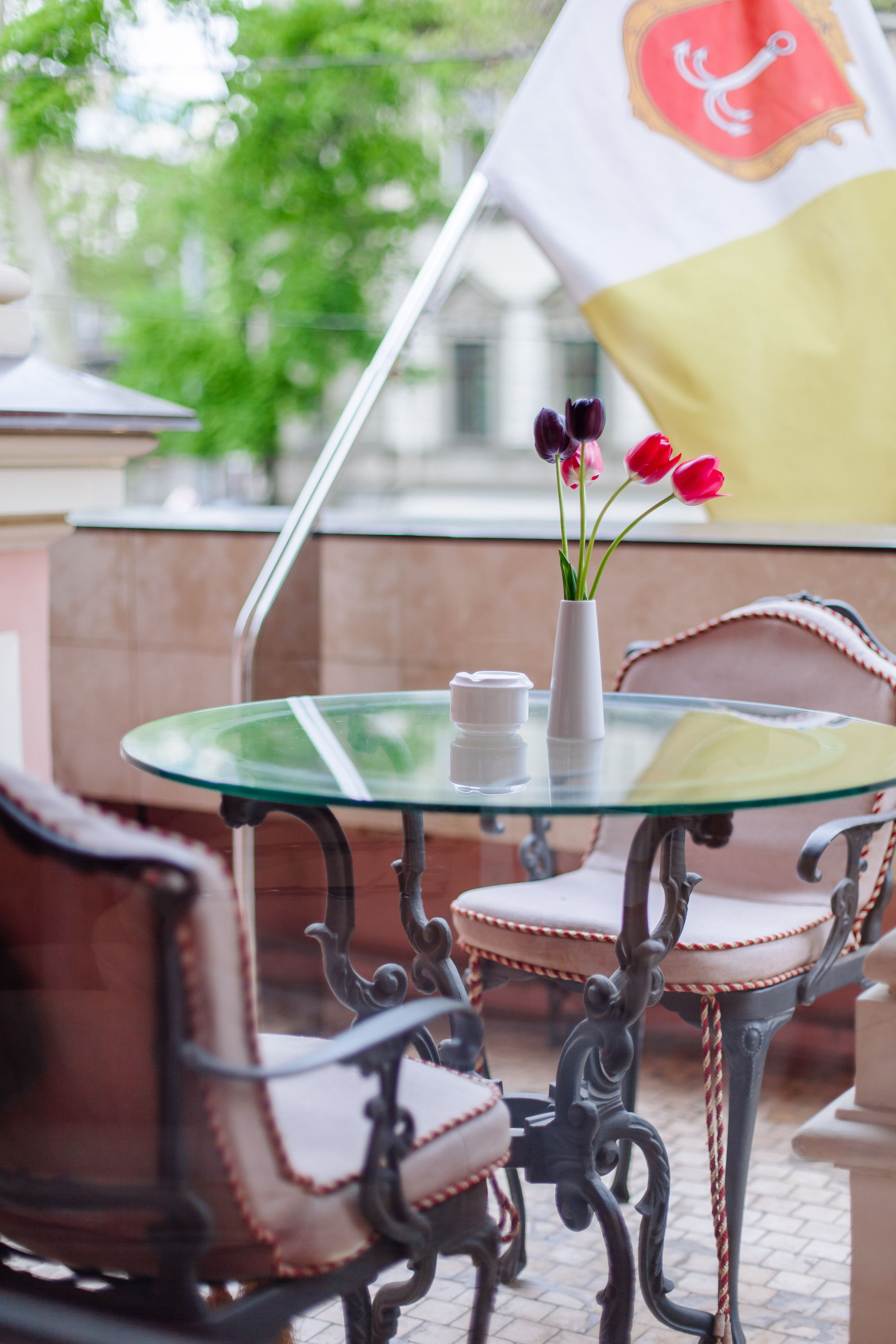 A round glass table with a white vase and red tulips, a flag, and ornate chairs.