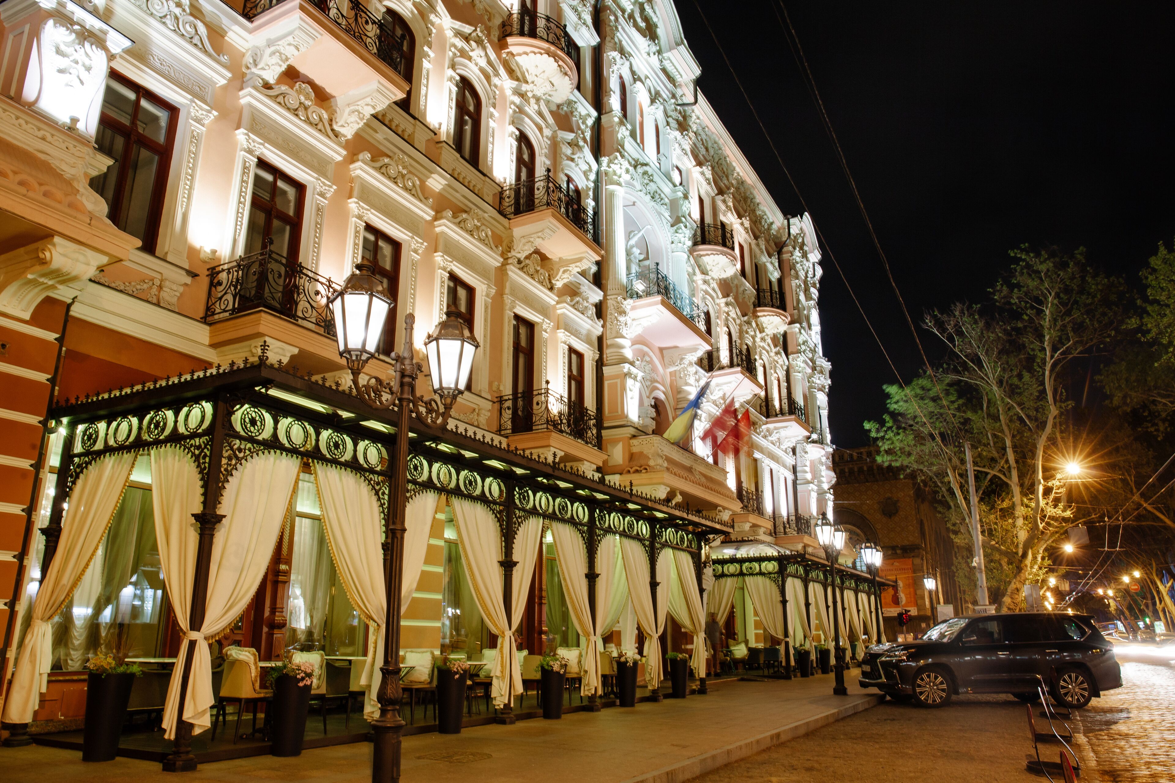 A historic building with ornate architecture, a covered outdoor seating area with tables and chairs, and a parked car on the street.
