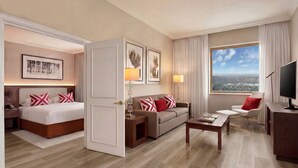 A hotel room with a bed, sofa, armchair, TV, and a large window with a city view. - NH Johannesburg Sandton Hotel (Johannesburg)