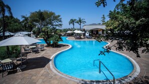 A swimming pool with lounge chairs and umbrellas, surrounded by palm trees and a building in the background. - NH Johannesburg Sandton Hotel (Johannesburg)