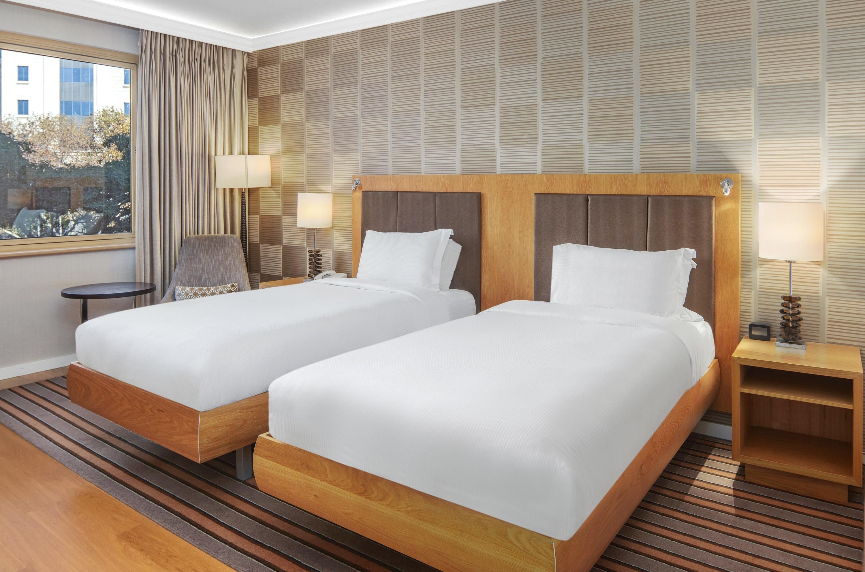 A hotel room with two beds, a window with curtains, a chair, a small table, and a lamp.
