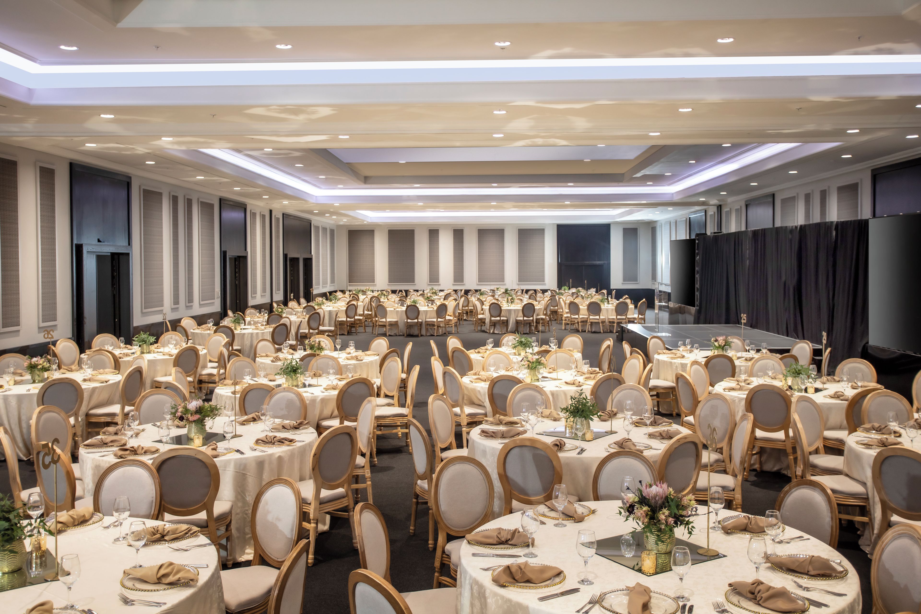 Une salle de banquet avec des tables et des chaises rondes, une scène et un grand écran.
