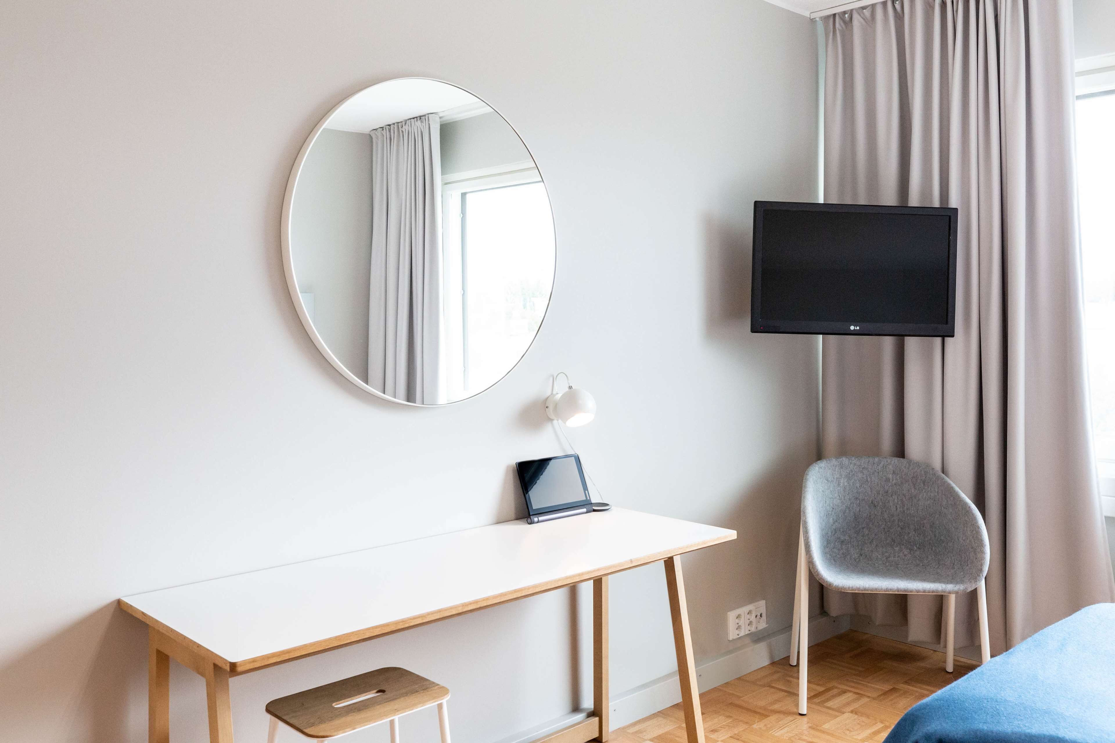 Habitación de hotel con escritorio blanco, una computadora portátil, una silla, televisor y espejo.