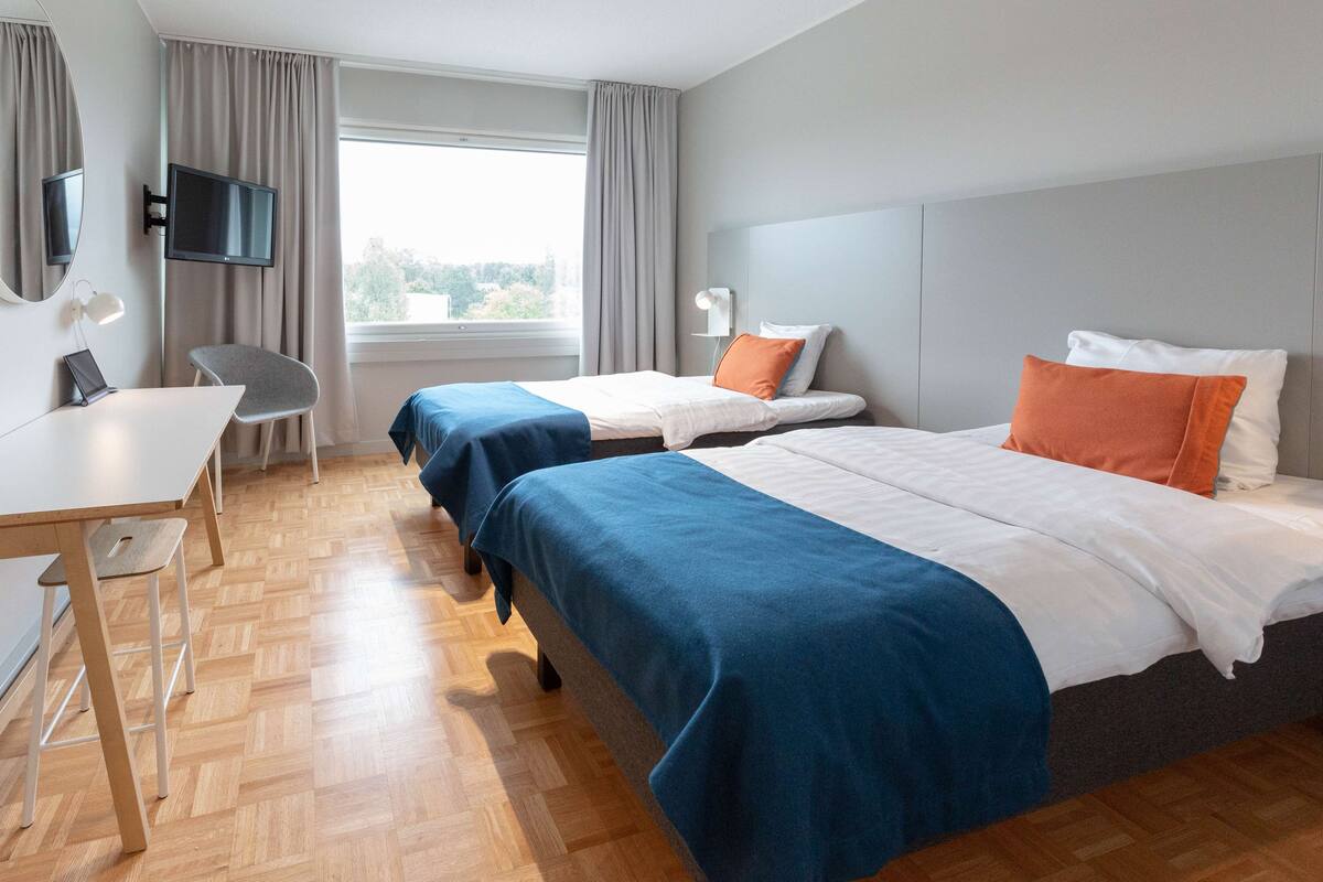 A hotel room with two beds, a desk, a chair, a TV, and a window with curtains.