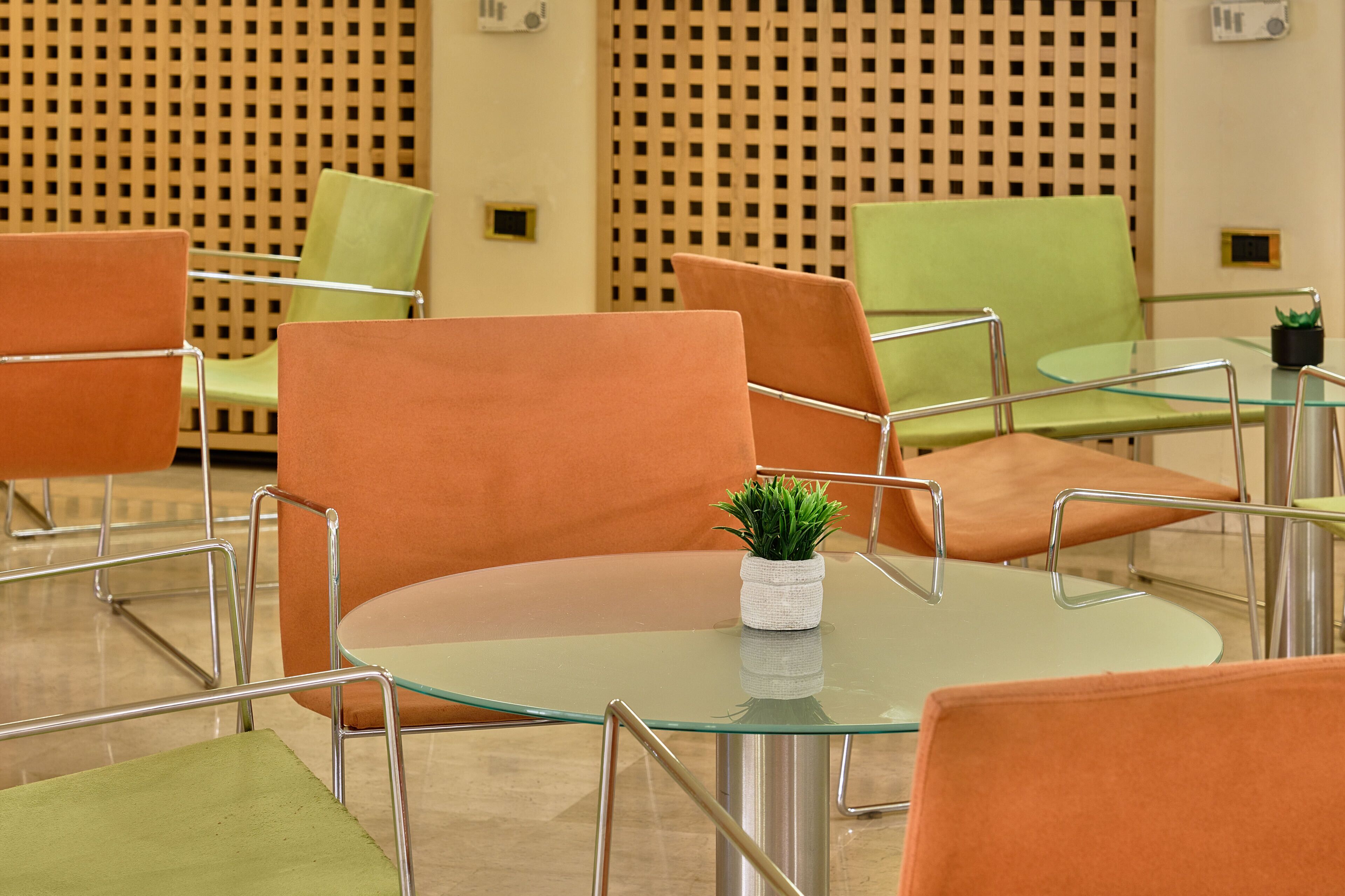 Un área de espera moderna con sillas anaranjadas y verdes, una mesa de vidrio y una pequeña planta.