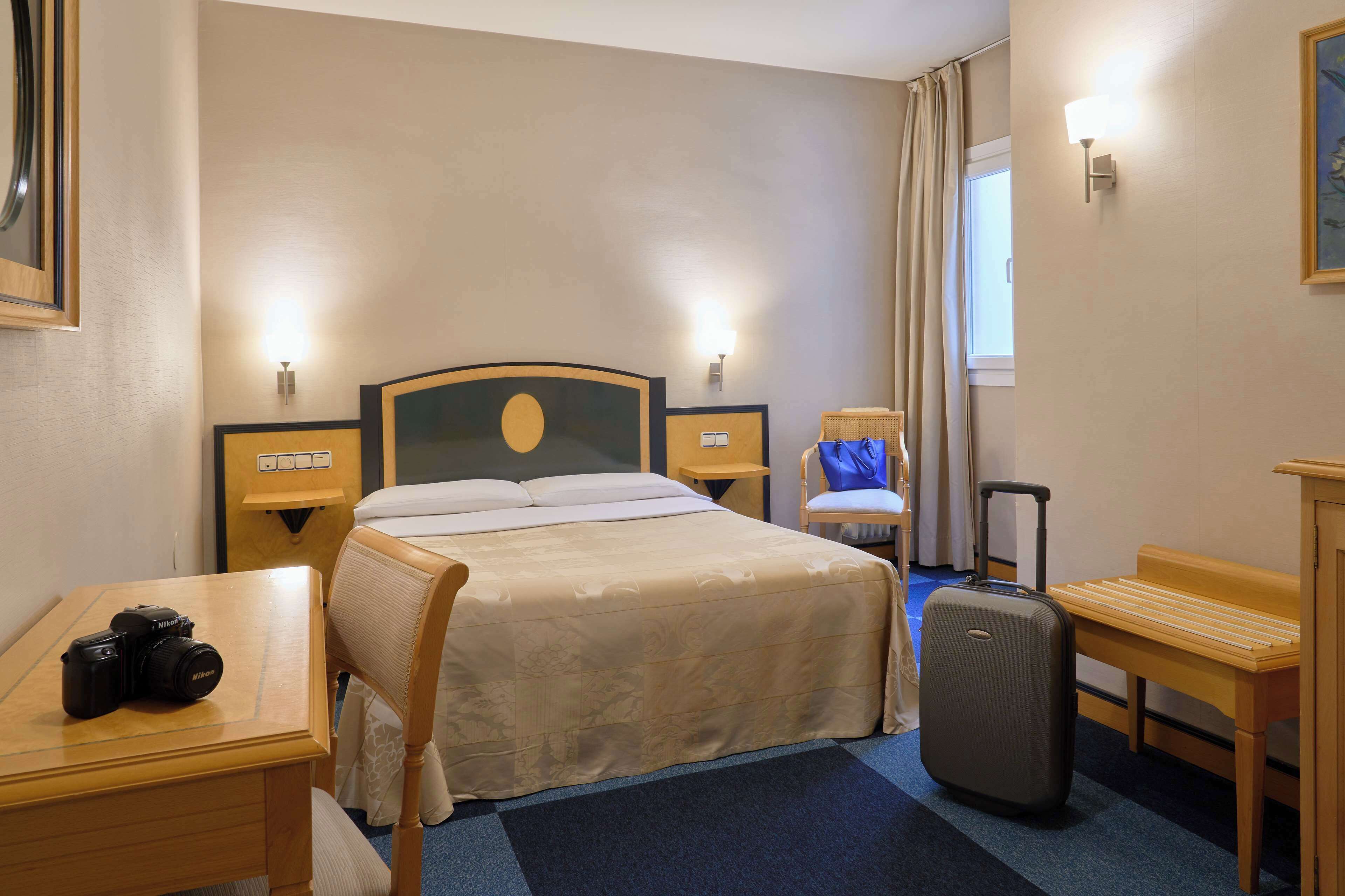 Une chambre d’hôtel avec un lit, un bureau, une chaise, une valise et une lampe.
