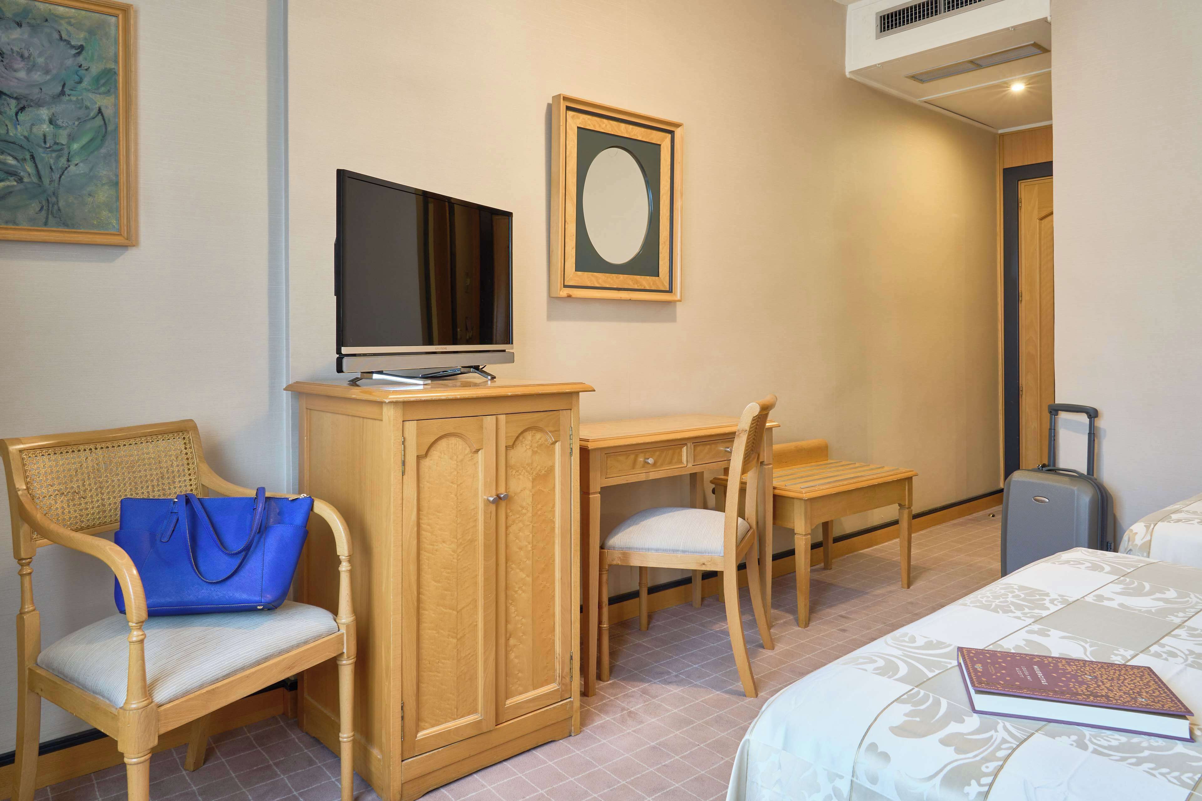 A hotel room with a wooden desk, a television, a chair with a blue bag, a small table, and a bed.