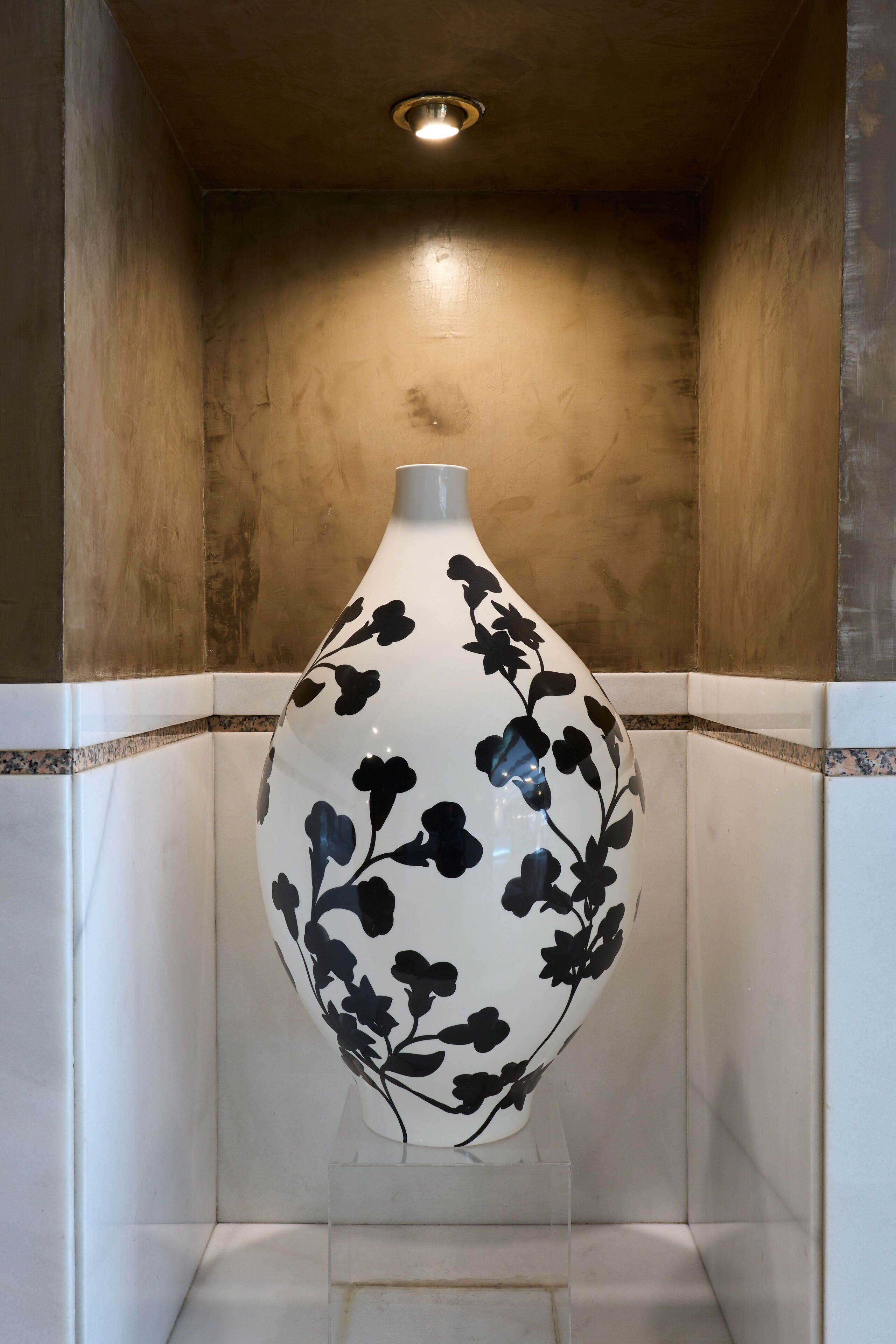 Un jarrón blanco grande con diseños florales negros sobre un pedestal transparente, en una esquina con paredes de azulejos y una lámpara empotrada en el techo.