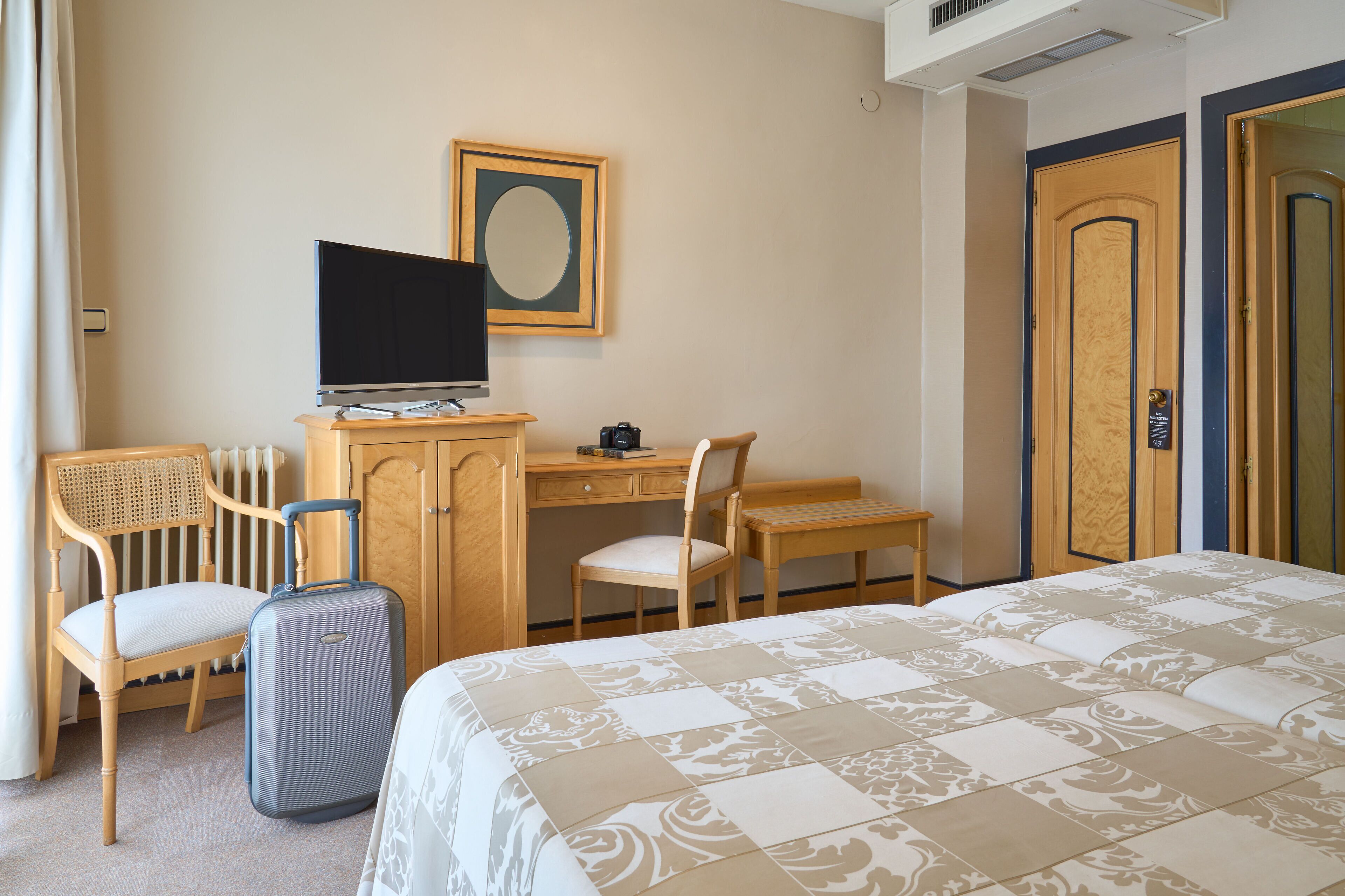 A hotel room with a bed, a desk with a chair, a television, and a suitcase.