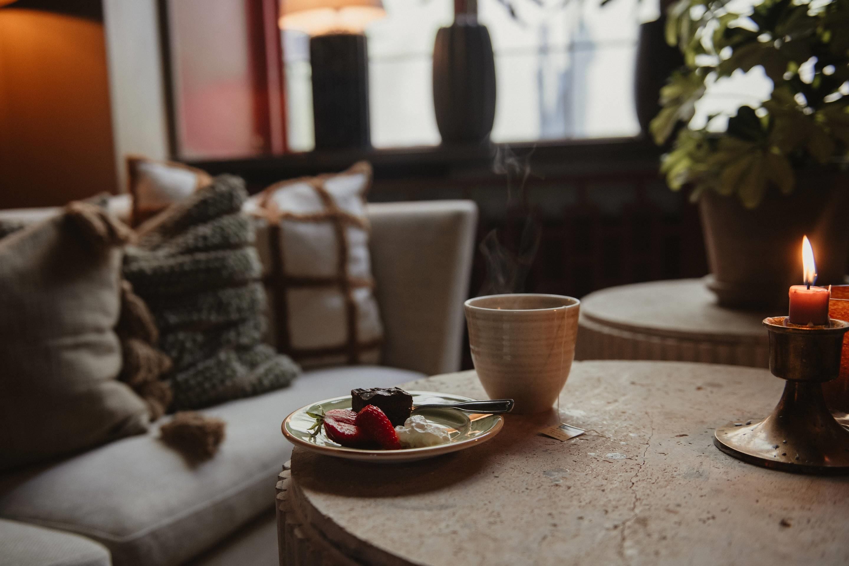 A cozy indoor setting with a round table, a candle, a plate with food, and a couch with cushions.
