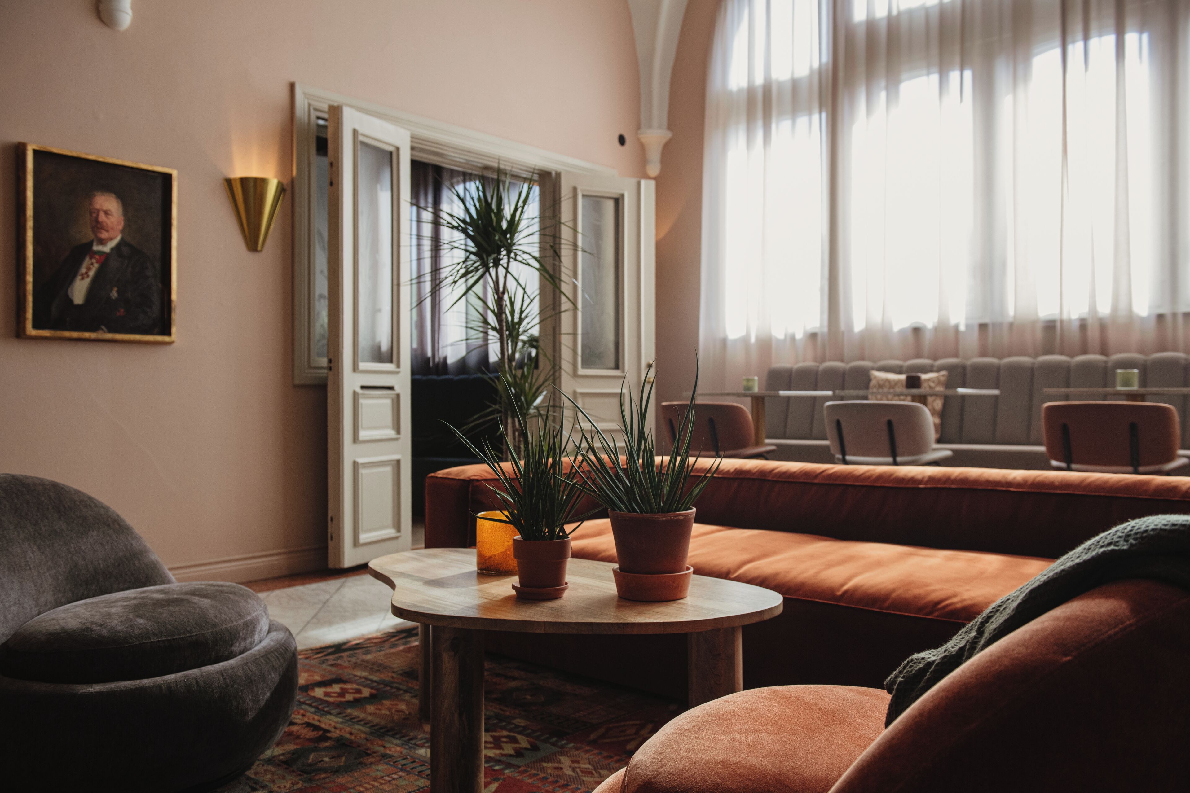 Ein gemütliches Wohnzimmer mit einer Couch, Sesseln, einem kleinen runden Tisch mit Topfpflanzen, einem Bild an der Wand und einem großen Fenster mit Vorhängen.