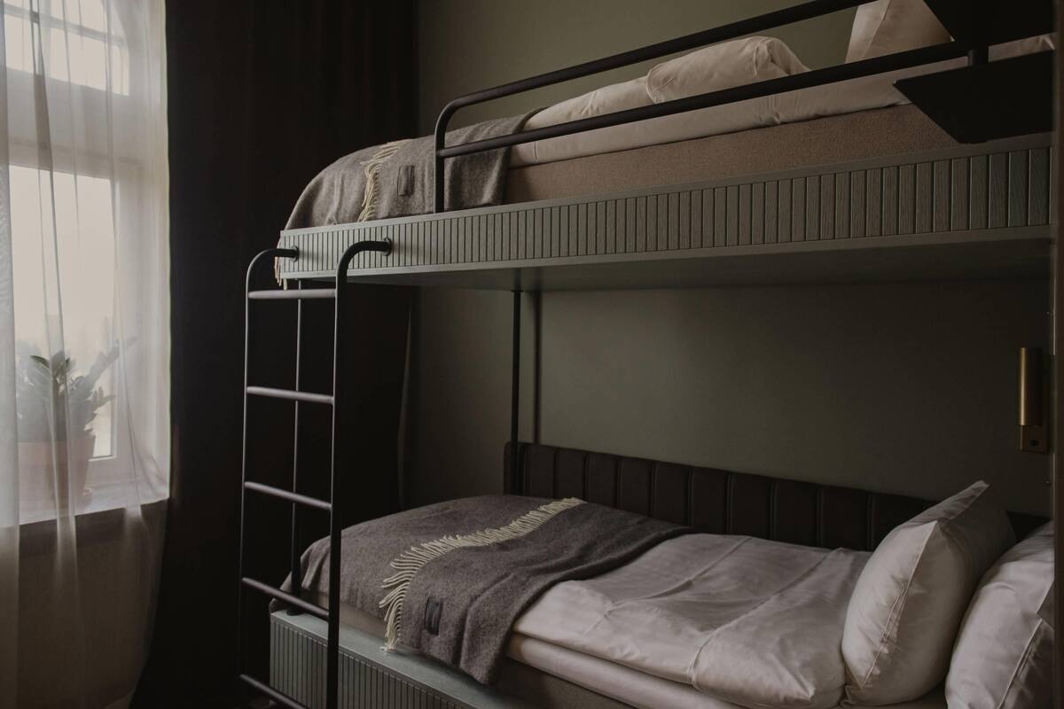 Uma cama beliche com escada, um radiador e uma janela com cortinas.
