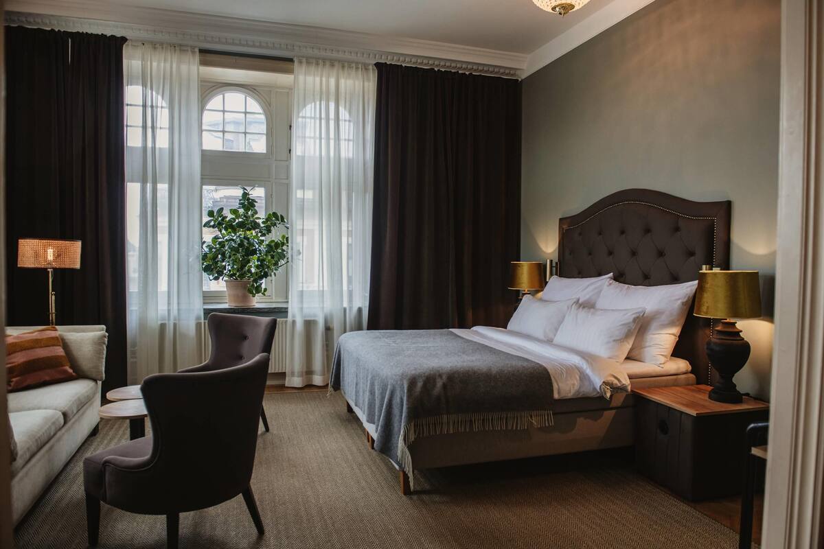 Quarto de hotel com uma cama grande, área de estar com sofá e poltronas, uma mesinha e uma planta em vaso.