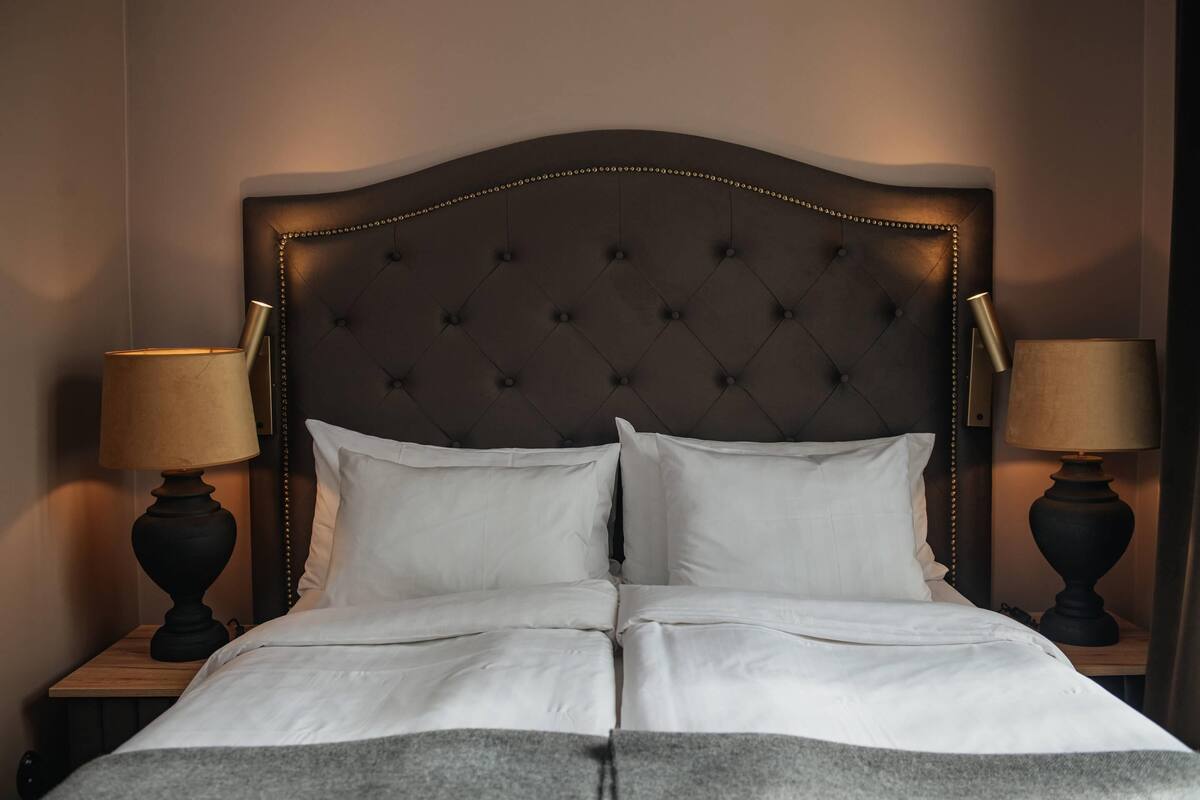 Cama bem arrumada com cabeceira estofada, duas luminárias de apoio e mesa de cabeceira de madeira.