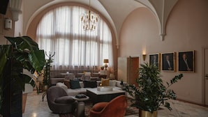A spacious living room with a high arched ceiling, a chandelier, and a large window with sheer curtains. There are several seating areas, including a sofa set and a coffee table. - Home Hotel Christian IV (Kristianstad)