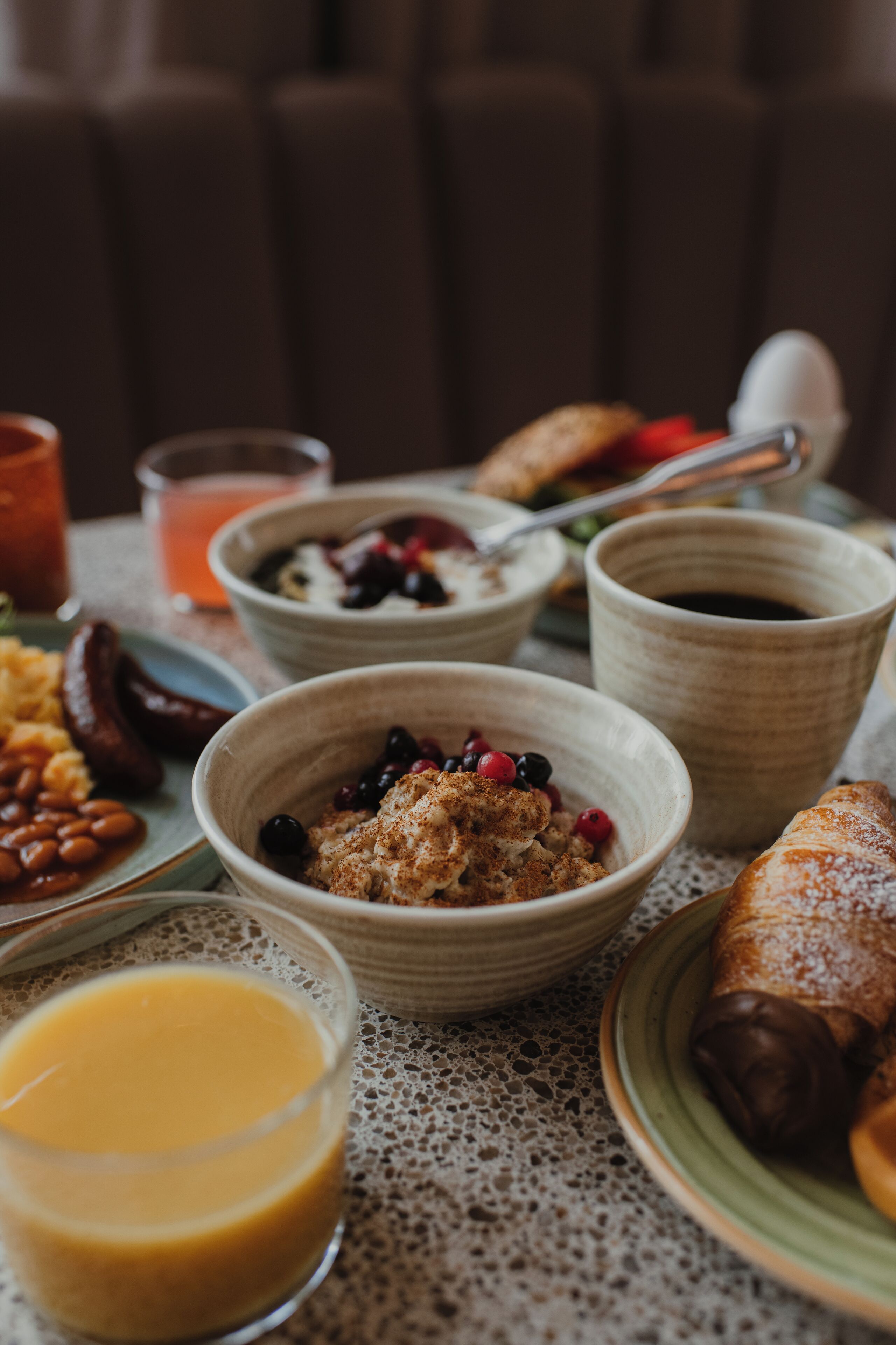 Una mesa de desayuno con diversos platos, entre ellos tazones de cereal, una bollería y un vaso de zumo de naranja.