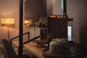 Una cafetera sobre una mesa de madera con una taza y platillo, una lámpara al fondo, y una silla.