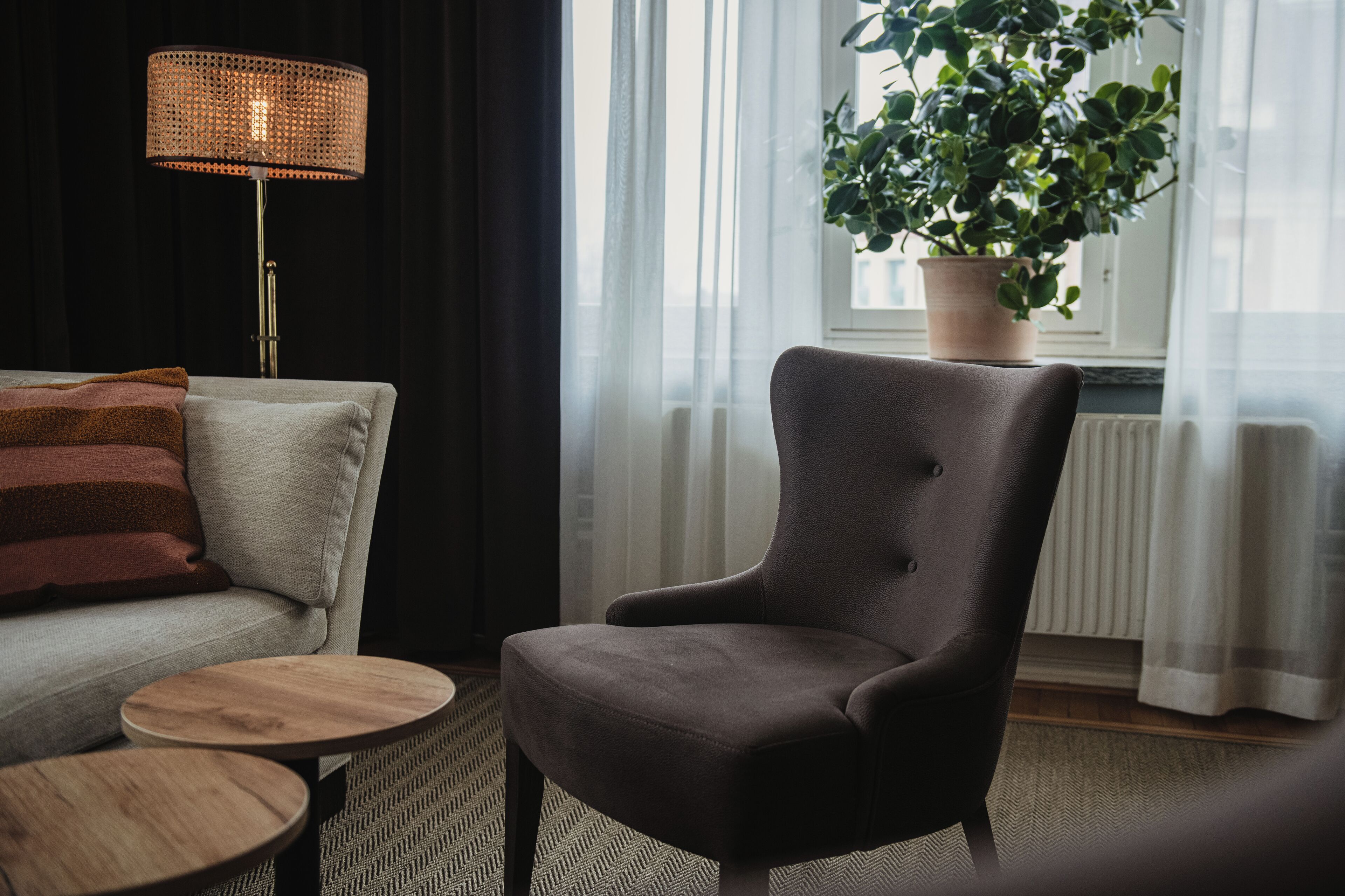 Ett vardagsrum med en soffa, en fåtölj, ett litet runt bord, en golvlampa och en krukväxt placerad vid ett fönster.