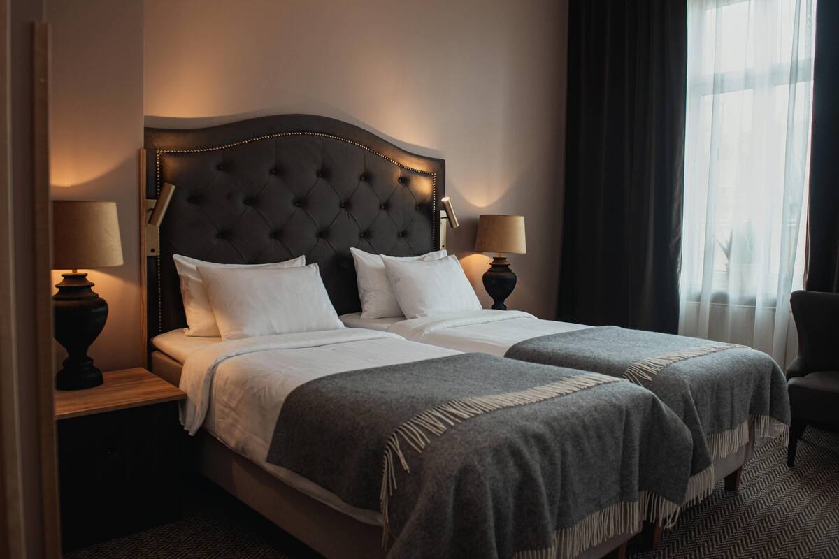 Uma cama de casal com cabeceira estofada, duas mesas de cabeceira com luminárias e vista para o exterior através de uma janela com cortinas leves.