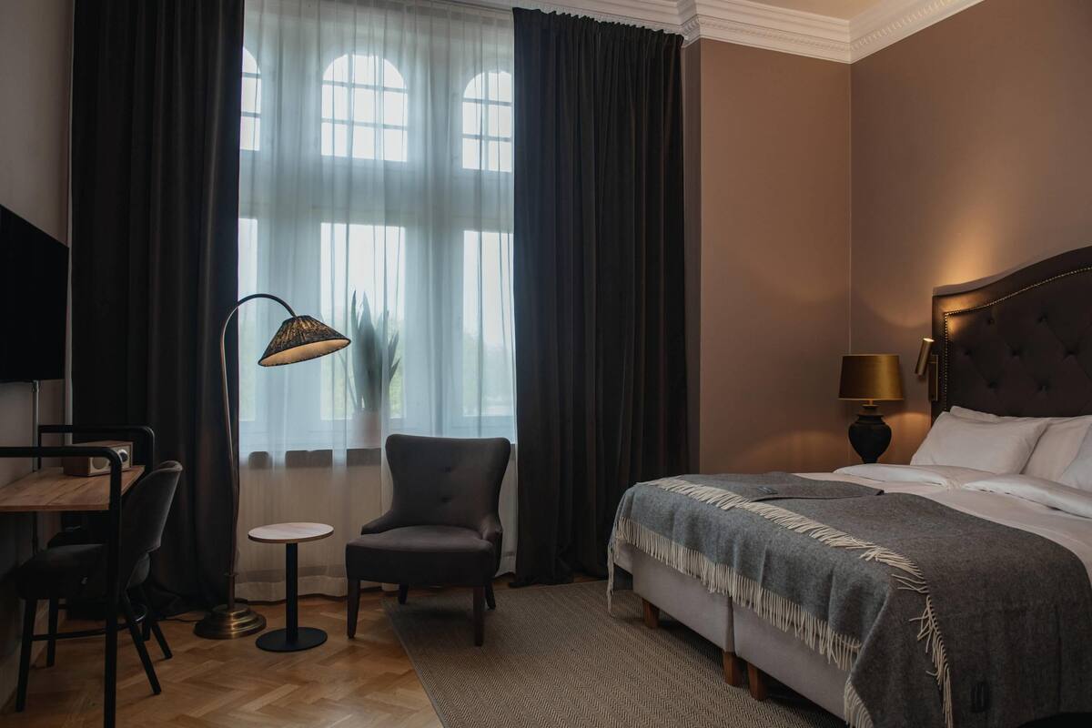 Quarto de hotel com cama, escrivaninha, cadeira, luminária e janela com cortinas.