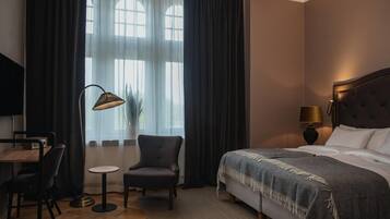 Ett hotellrum med en säng, ett skrivbord, en stol, en lampa och ett fönster med gardiner.