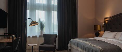 Une chambre d’hôtel avec un lit, un bureau, une chaise, une lampe et une fenêtre avec des rideaux.