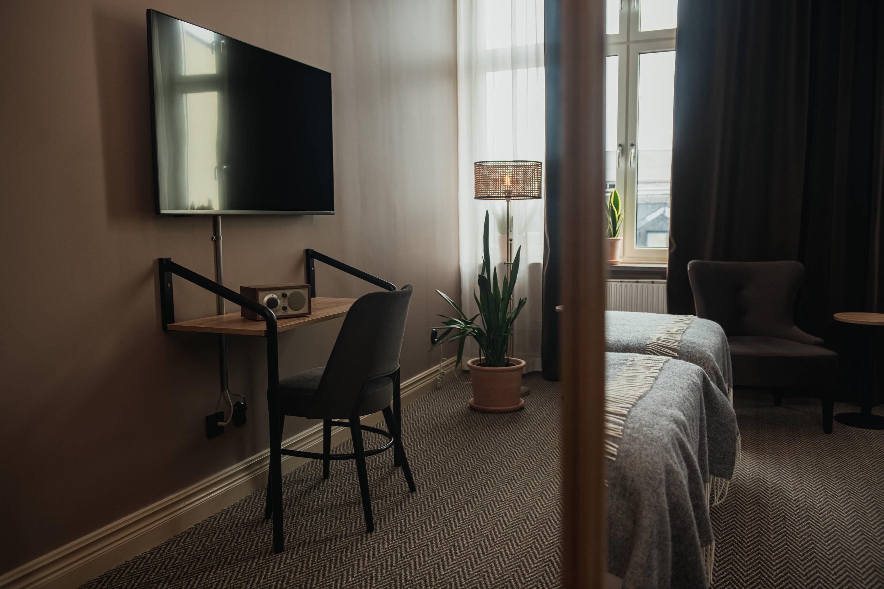 Una habitación de hotel con cama, escritorio y silla, televisor montado en la pared, planta en maceta y ventana con cortinas.