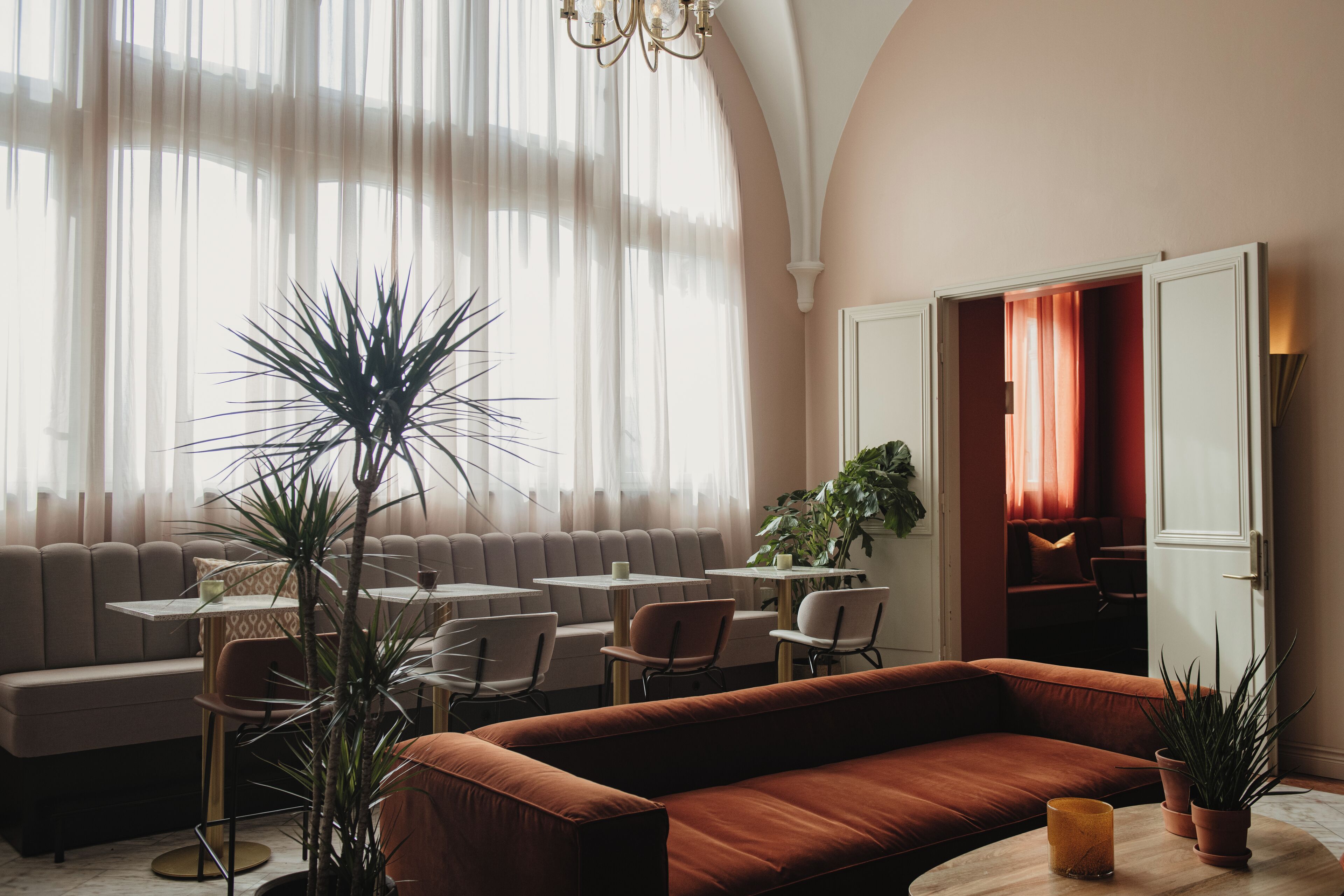 A modern lounge with a sofa, chairs, and tables, featuring large windows and potted plants.