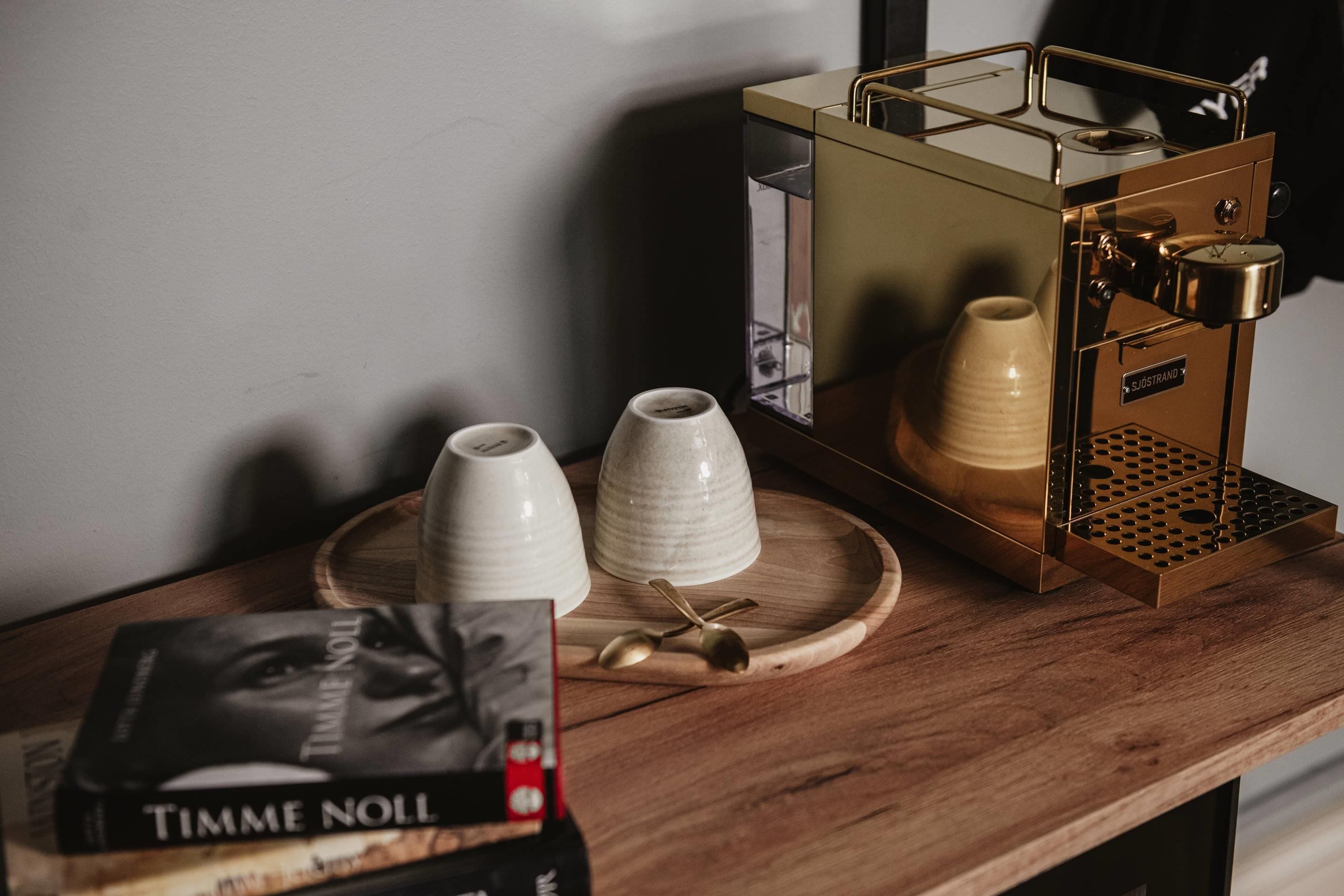 A coffee machine on a wooden table with two white cups and a spoon.