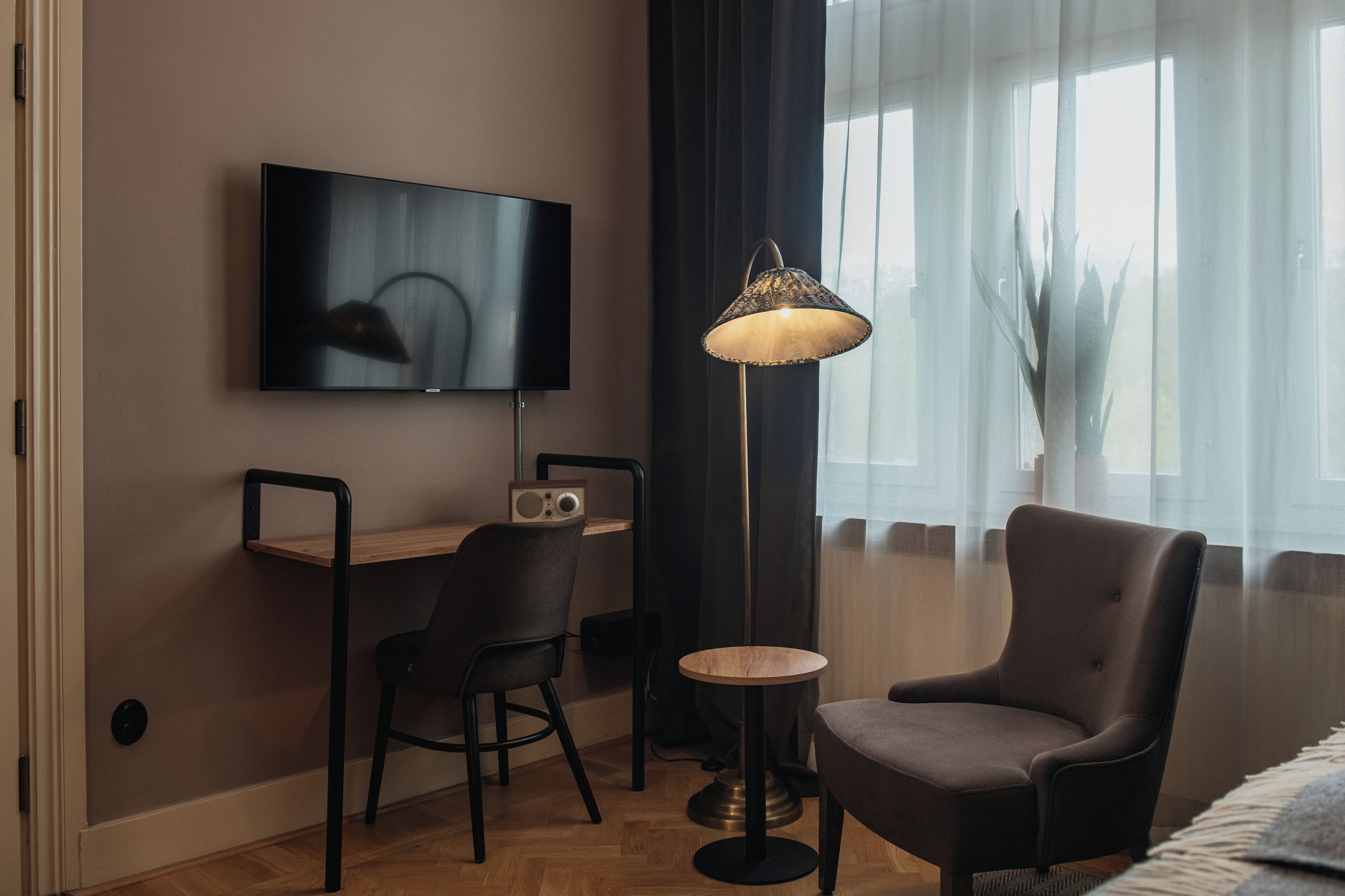 Ett modernt hotellrum med en platt-TV, ett skrivbord med en stol, ett runt bord och en golvlampa.