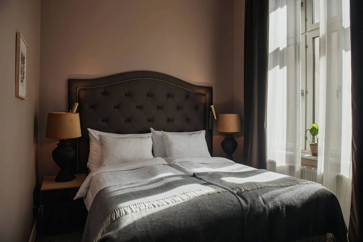Cama bem arrumada com cabeceira estofada, duas luminárias de apoio e uma janela com cortinas.