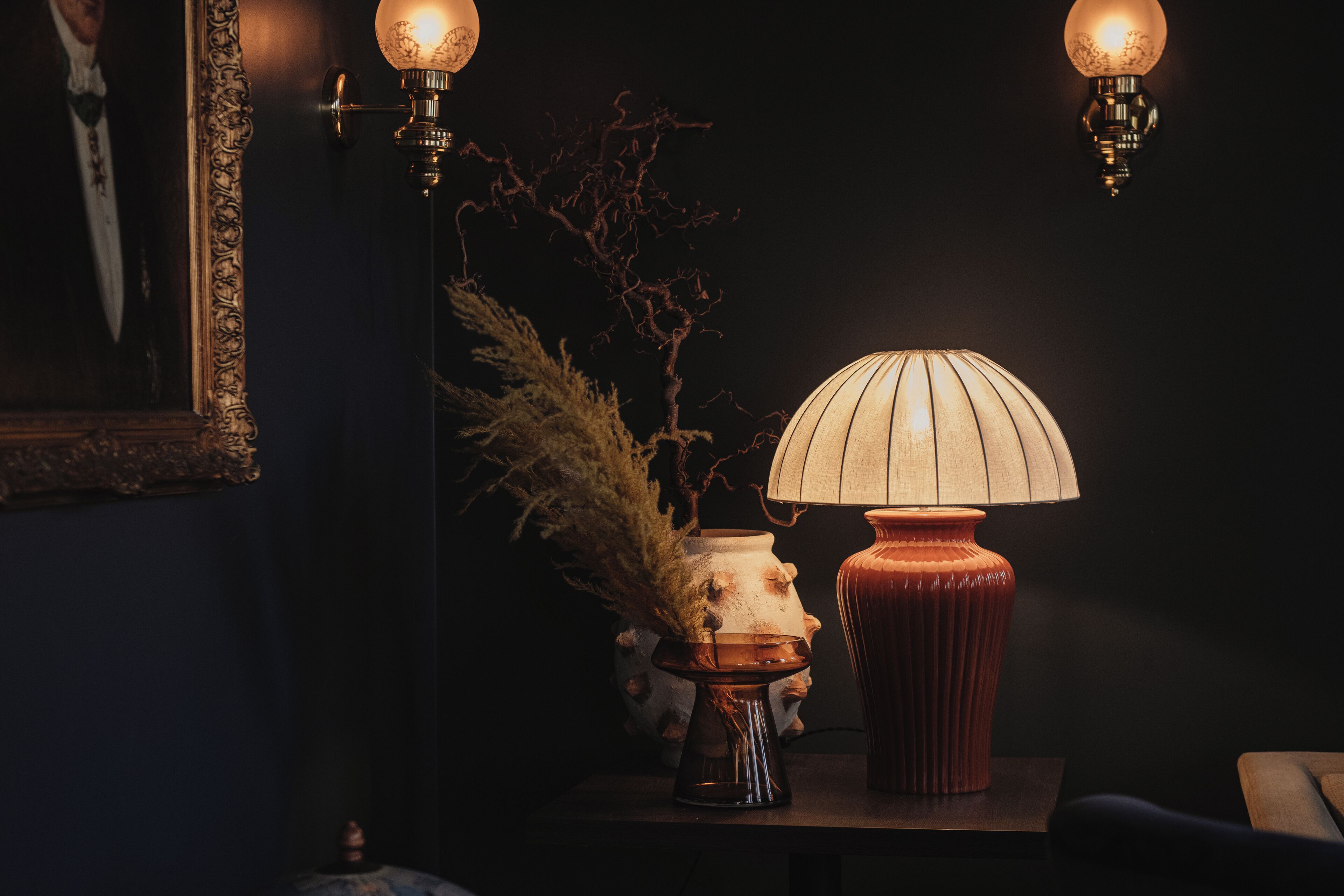 Una habitación oscura con una mesa que tiene una lámpara, un florero con plantas secas y un cuadro enmarcado.