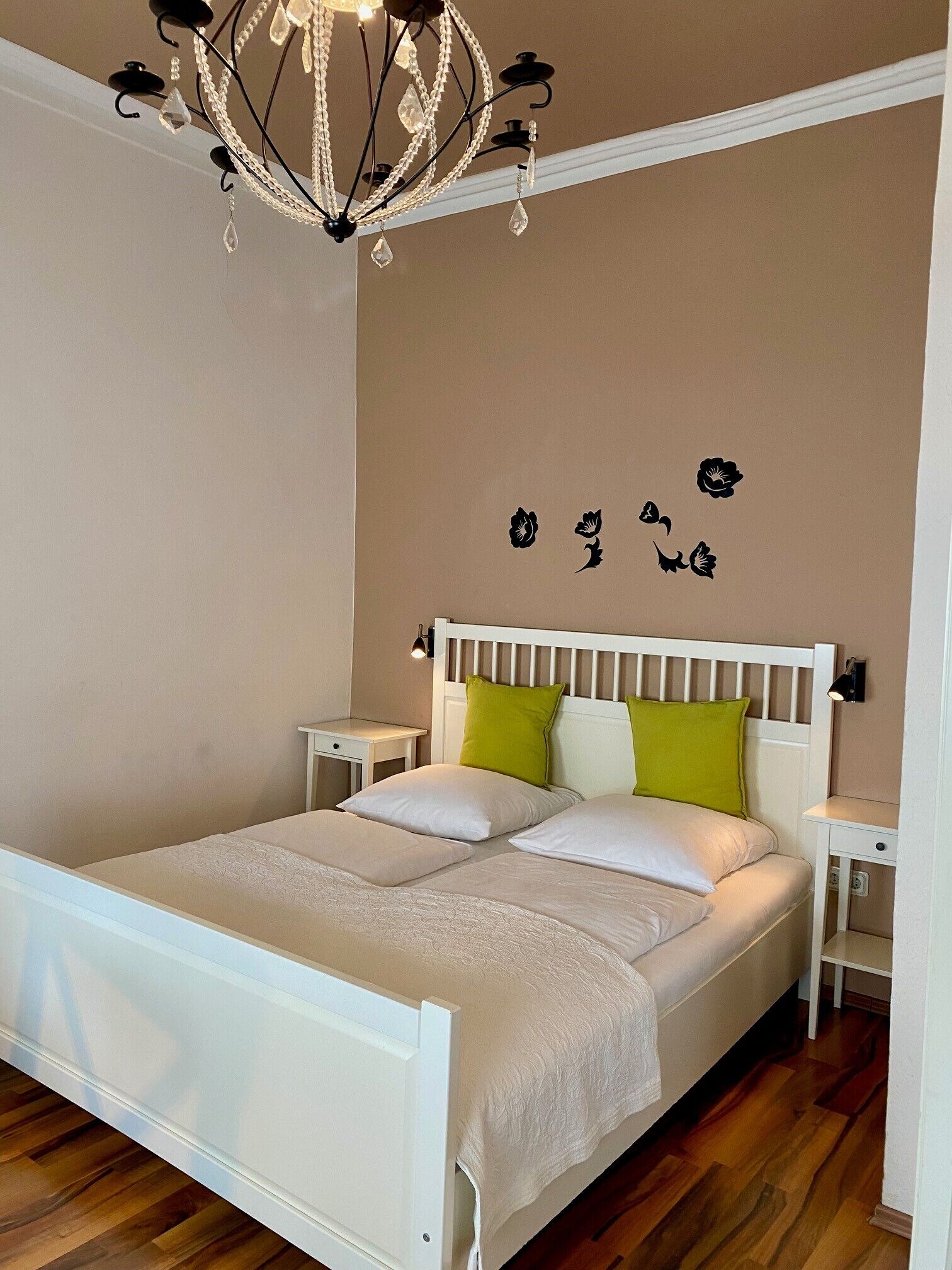 Un dormitorio con una cama blanca, almohadas verdes y blancas, una mesita de noche blanca, una lámpara de pared y un candelabro.