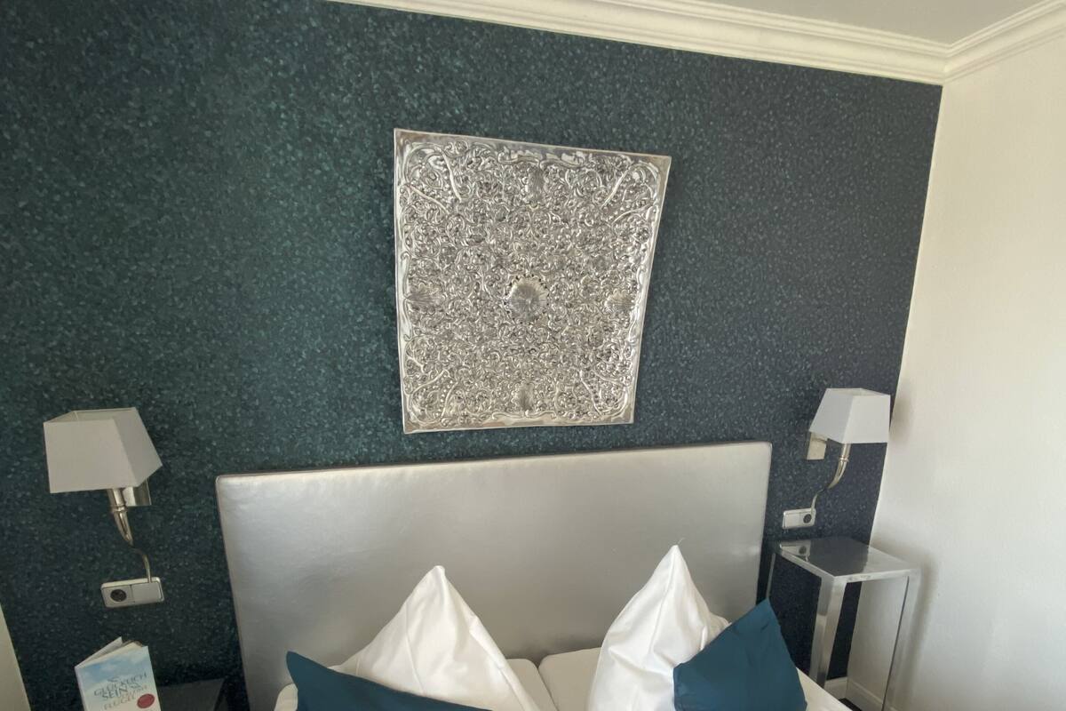 A hotel room with a neatly made bed, two pillows, a bedside table with a lamp, and a decorative wall piece.